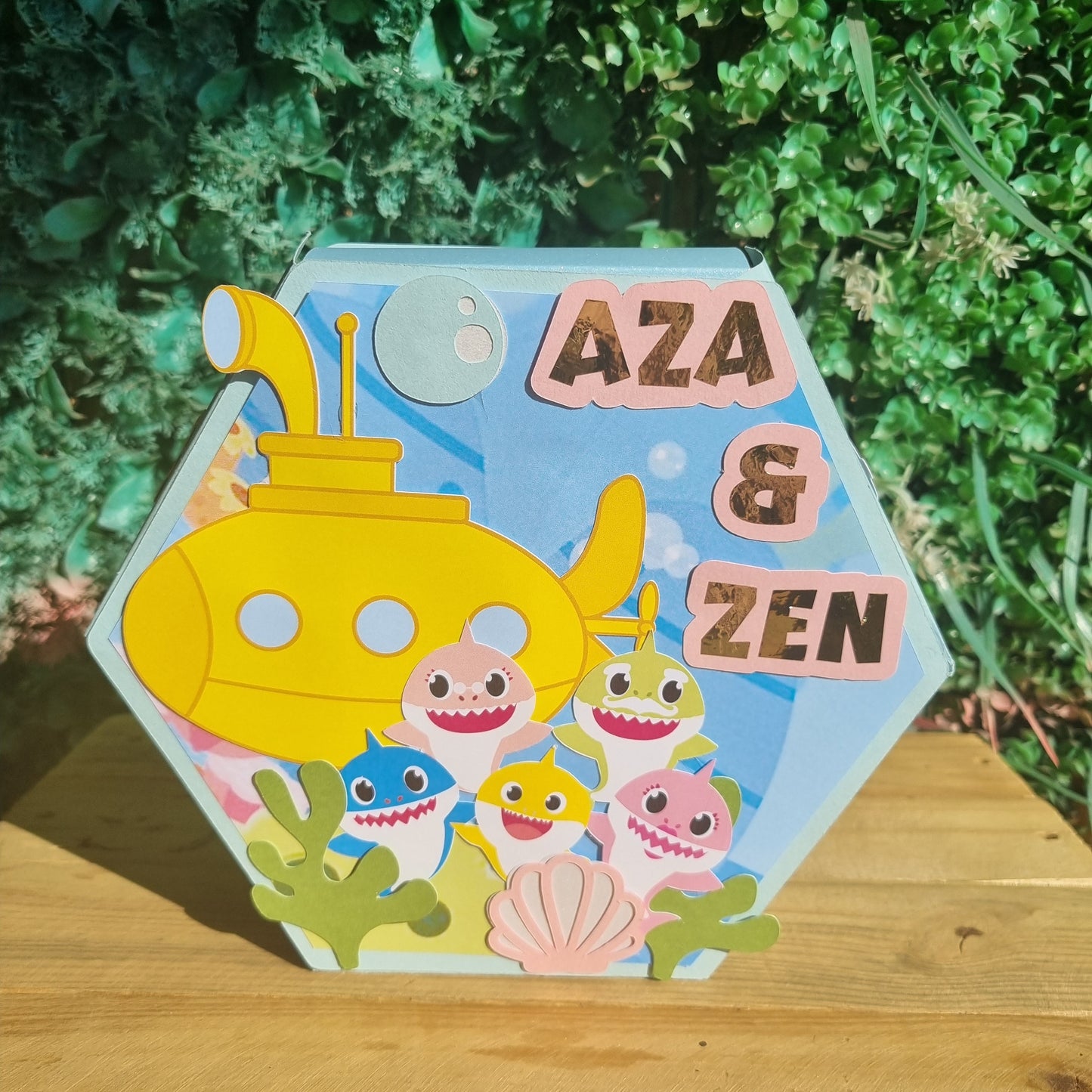 Colorful children's book titled 'Aza & Zen' with a cartoon submarine on a wooden surface and green foliage background.