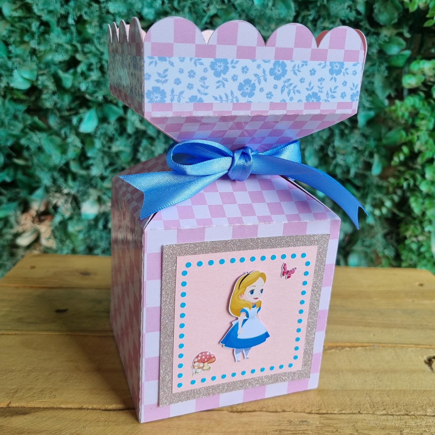 Pink checkered gift box with a blue ribbon on a wooden surface with greenery in the background