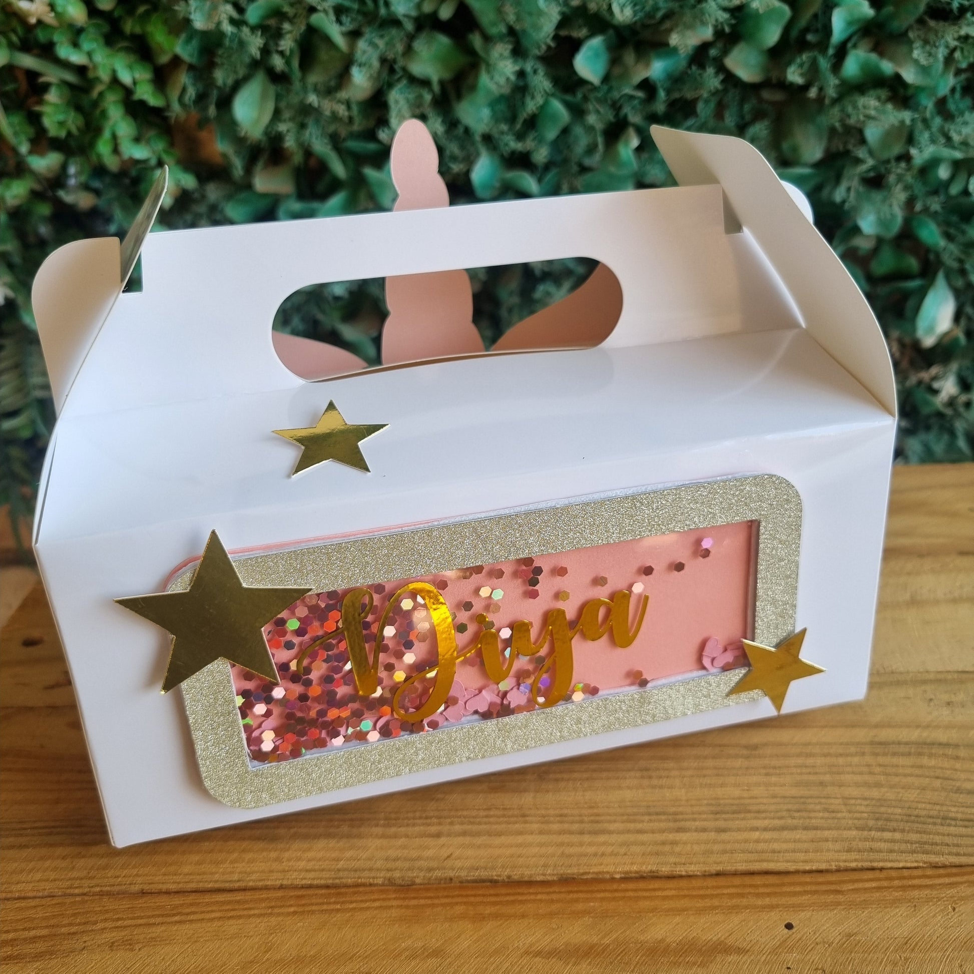 White cake box with gold stars and a pink cake inside on a wooden surface.