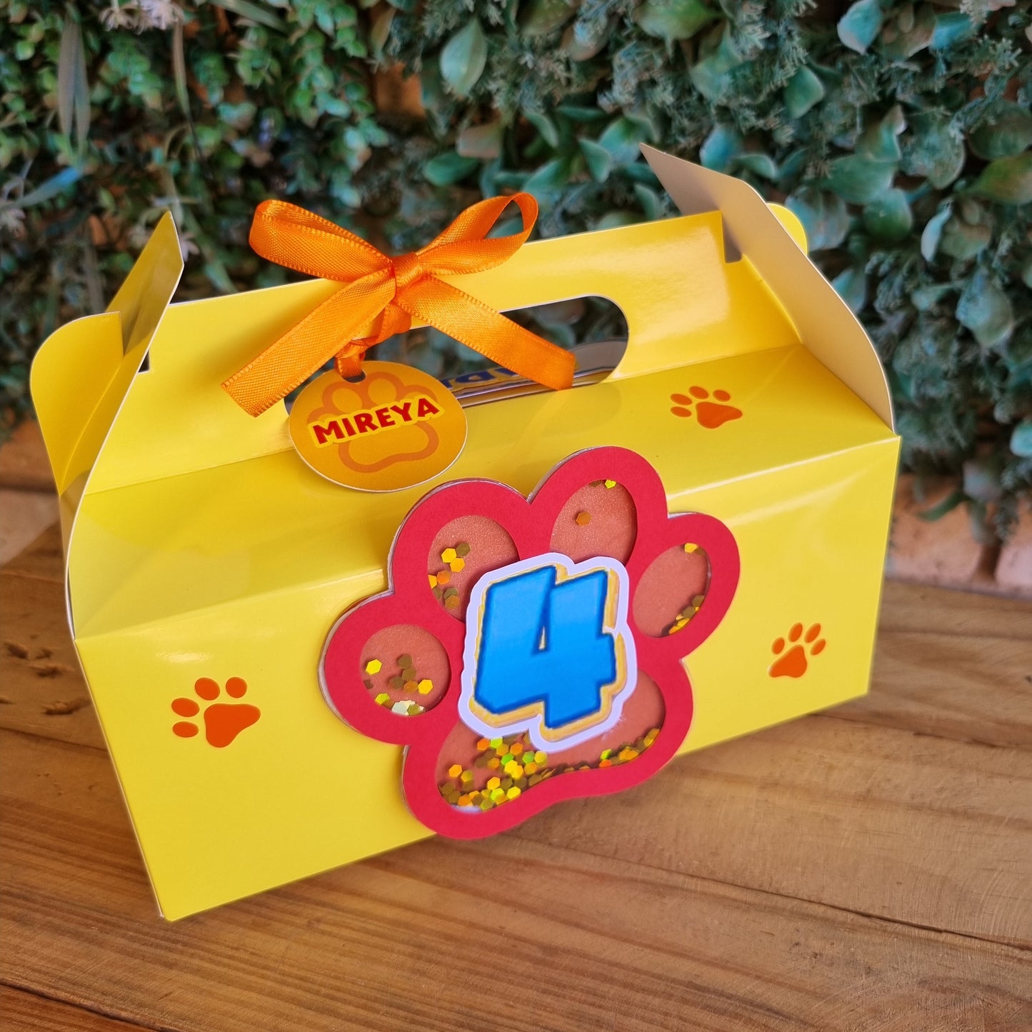 Yellow gift bag with a decorative flower and bow, featuring the brand 'Mireya', against a wooden and green leafy background.