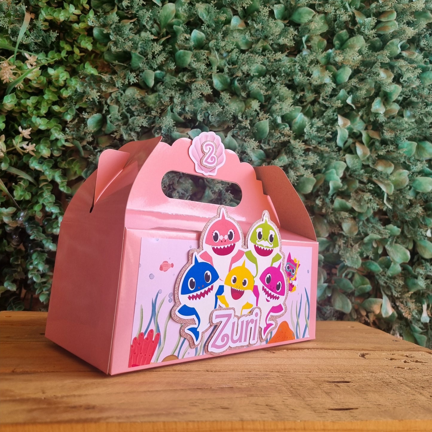 Pink children's gift box with colorful design on a wooden surface and green foliage background