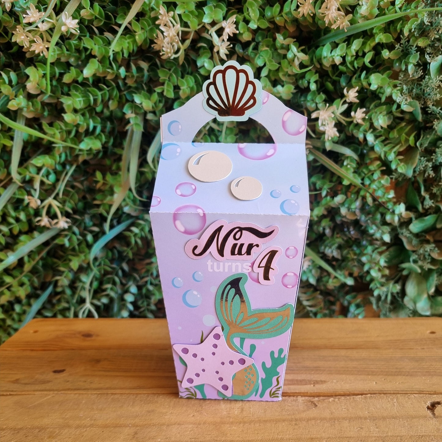 Decorative paper party box with mermaid design on a wooden surface and greenery background