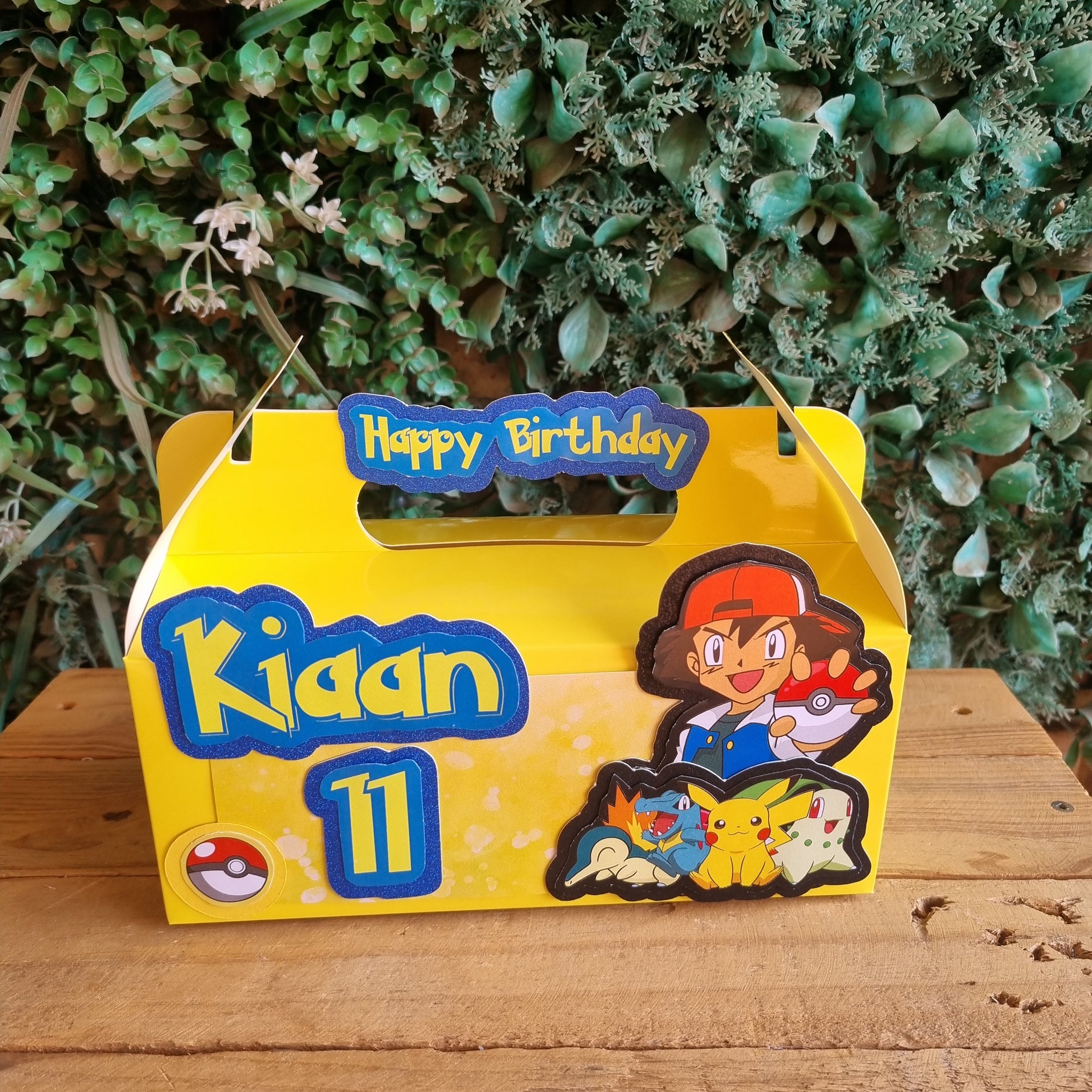 Yellow 'Happy Birthday' card with cartoon characters on a wooden surface and green plants.