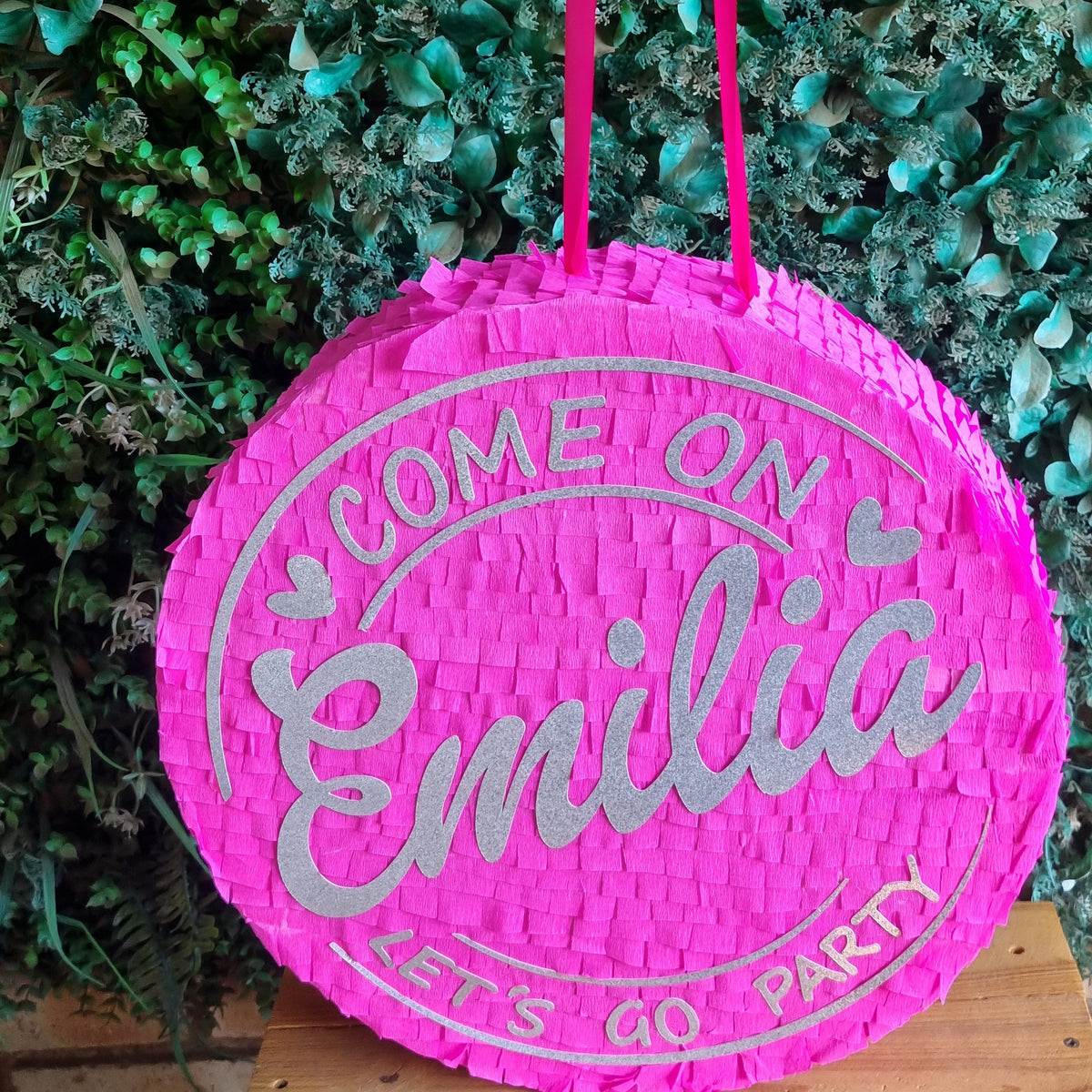 A bright pink woven round Barbie piñata bag with silver glitter text reading 'COME ON Emilia LET'S GO PARTY', hanging against a green leafy background.