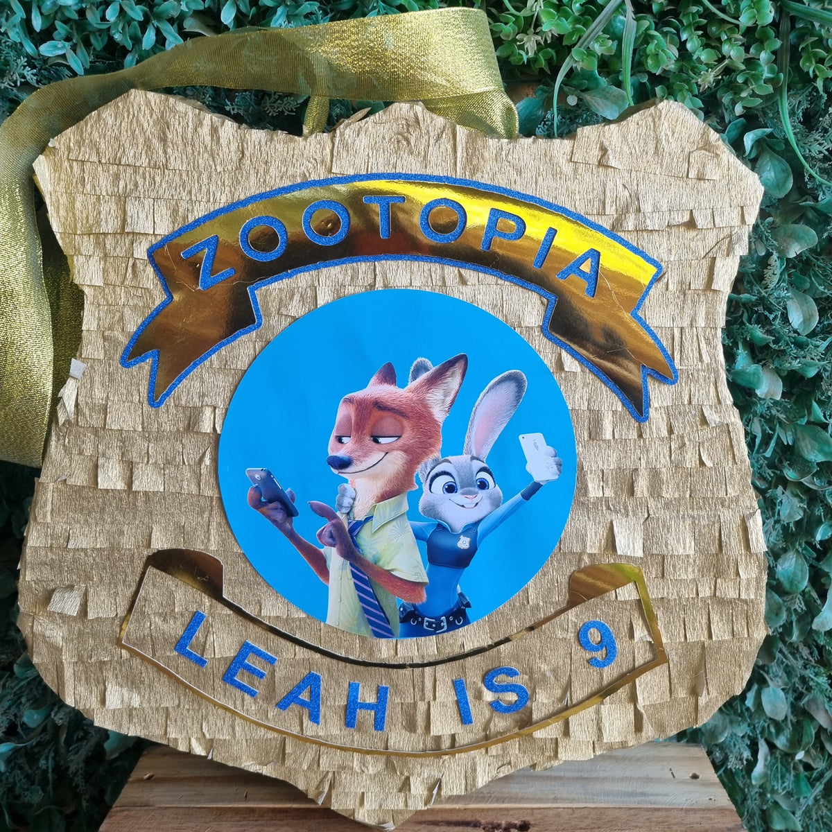 A custom Zootopia-themed wooden pinata shaped like a police badge, featuring the characters Judy Hopps and Nick Wilde on a blue circle, with 'ZOOTOPIA' and 'LEAH IS 9' in blue lettering. The pinata is decorated with a gold ribbon and hangs against a green hedge background.
