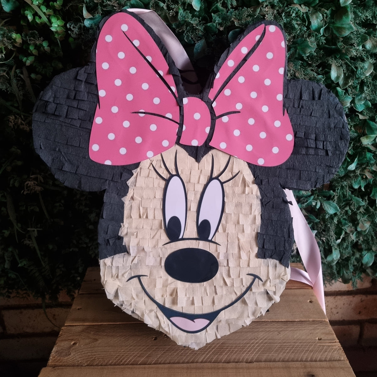 A Minnie Mouse-themed pinata shaped like her face, featuring a pink polka-dot bow, black ears, and a natural wood-textured face, displayed against a green leafy background.