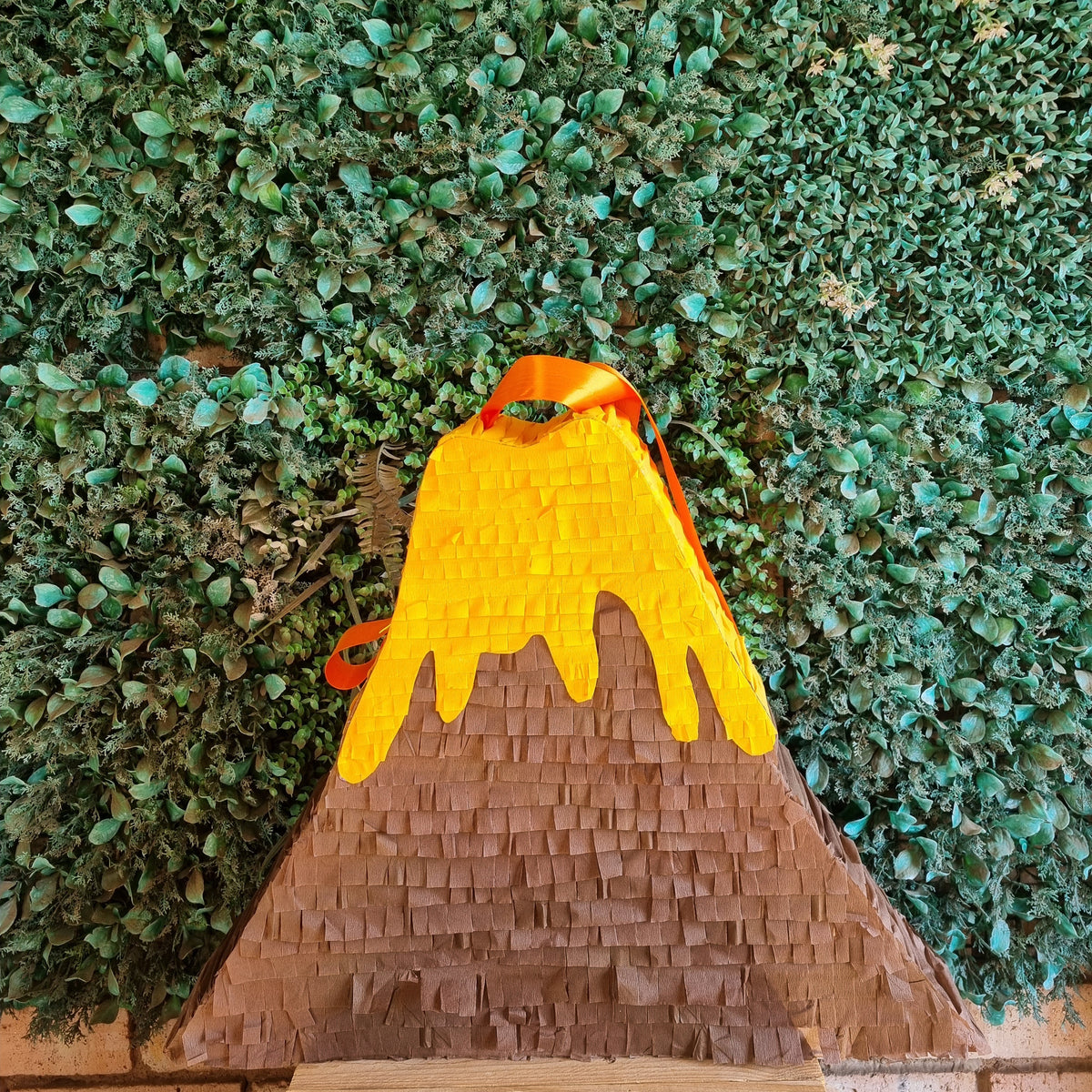 A volcano-shaped pinata with a textured brown base, dripping yellow lava, and orange-red accents, photographed against a green leafy hedge background.