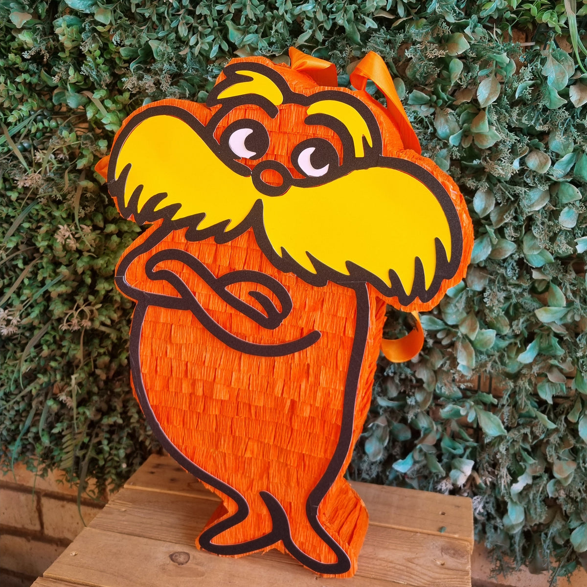 A Dr. Seuss Lorax pinata shaped like the character, standing on a wooden surface against a green leafy hedge background.