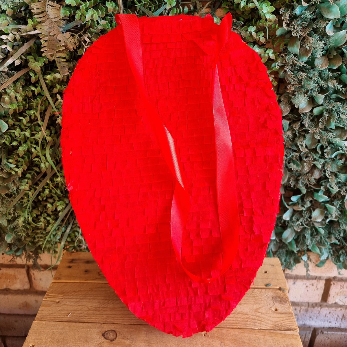 A bright red woven tote bag with two long handles, displayed on a wooden surface in front of a green leafy wall.