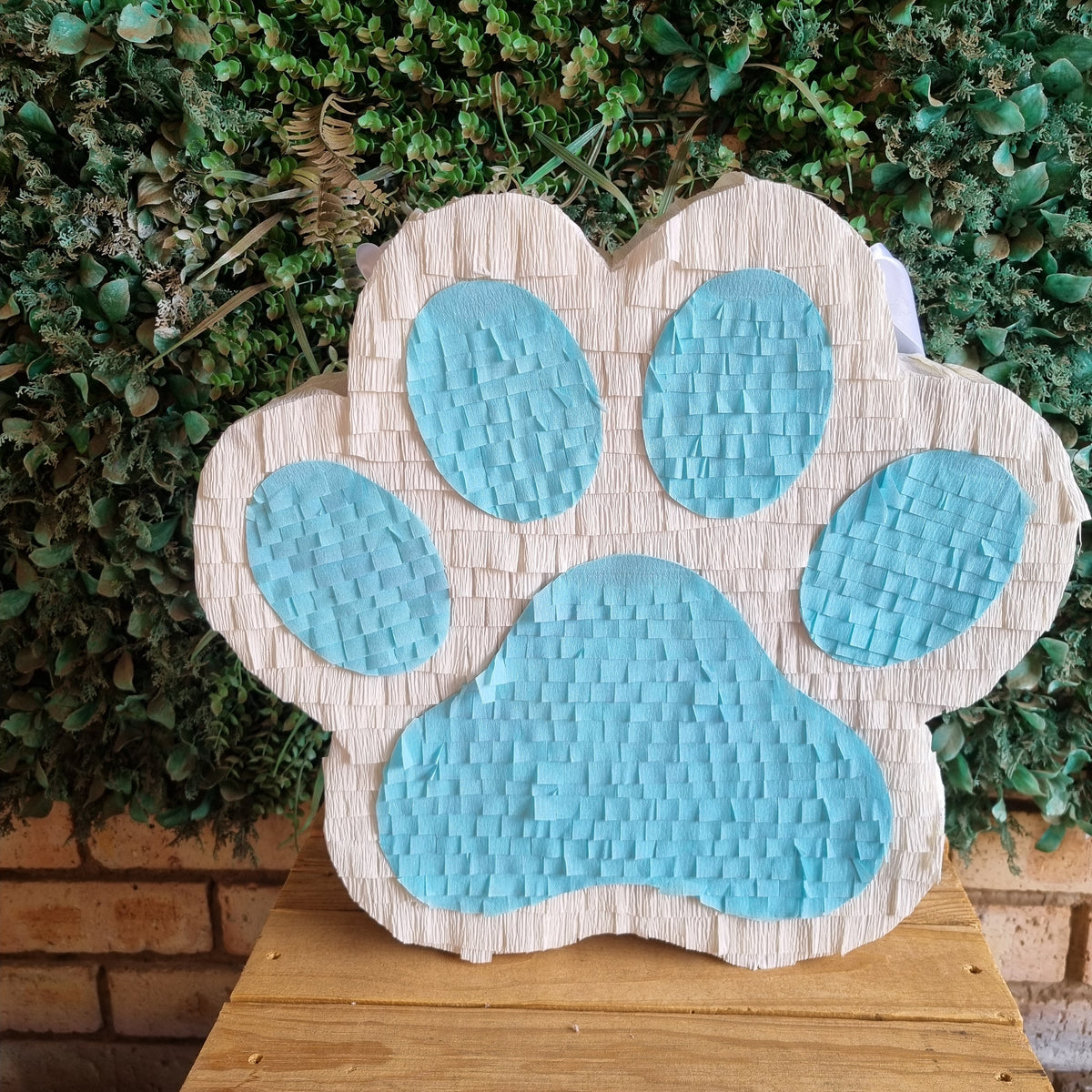 A paw print shaped pinata with a textured beige paper body and light blue accents, displayed on a wooden table in front of a green leafy wall.