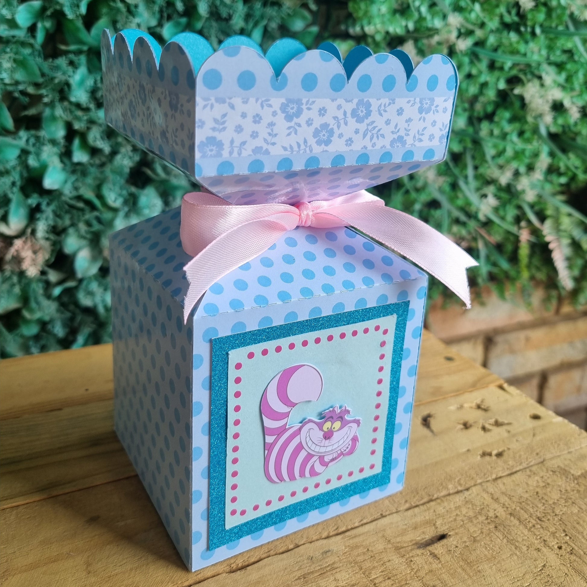 Decorative box with a pink ribbon on a wooden surface with greenery in the background