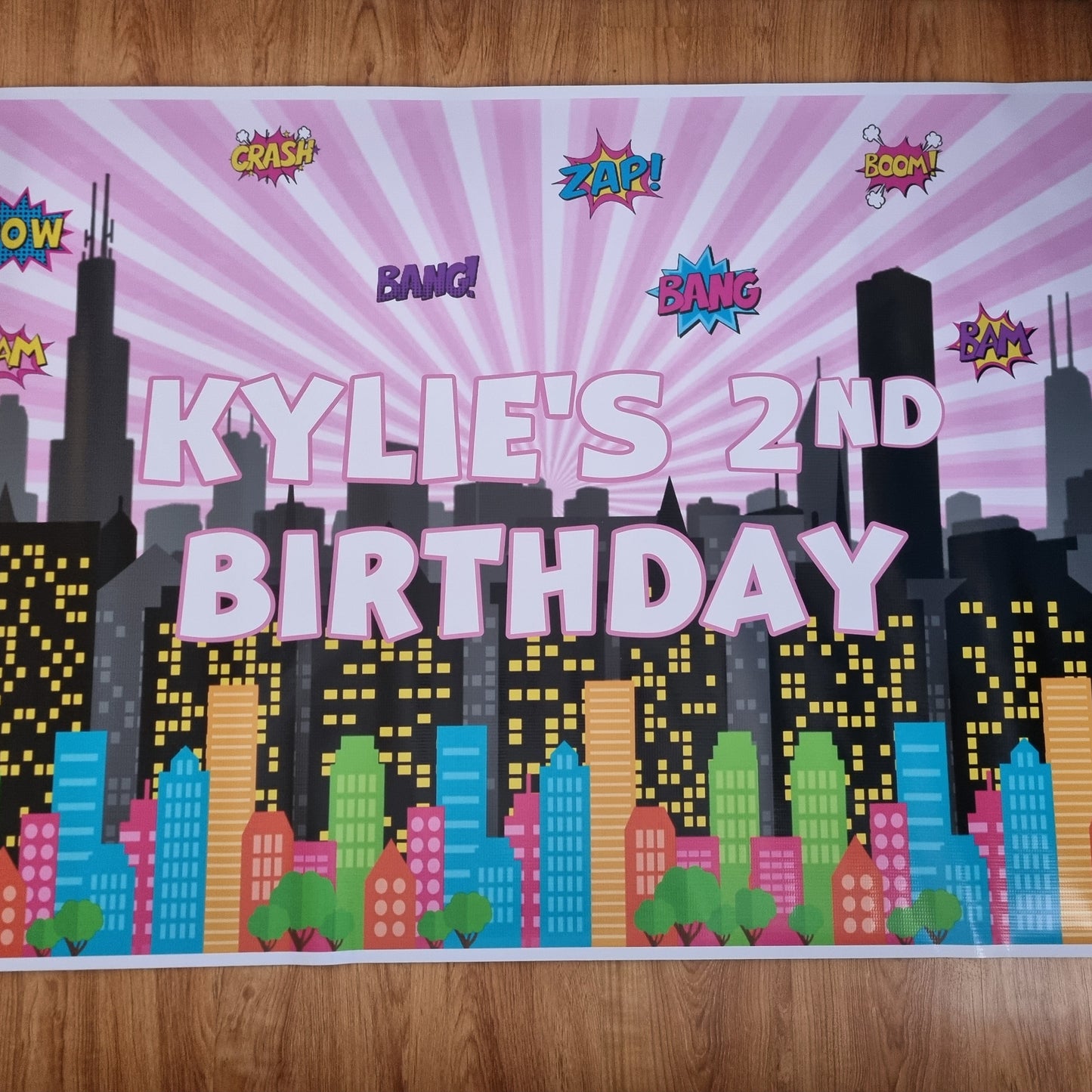 Birthday banner with cityscape design and text 'Kylie's 2nd Birthday' on a wooden floor.