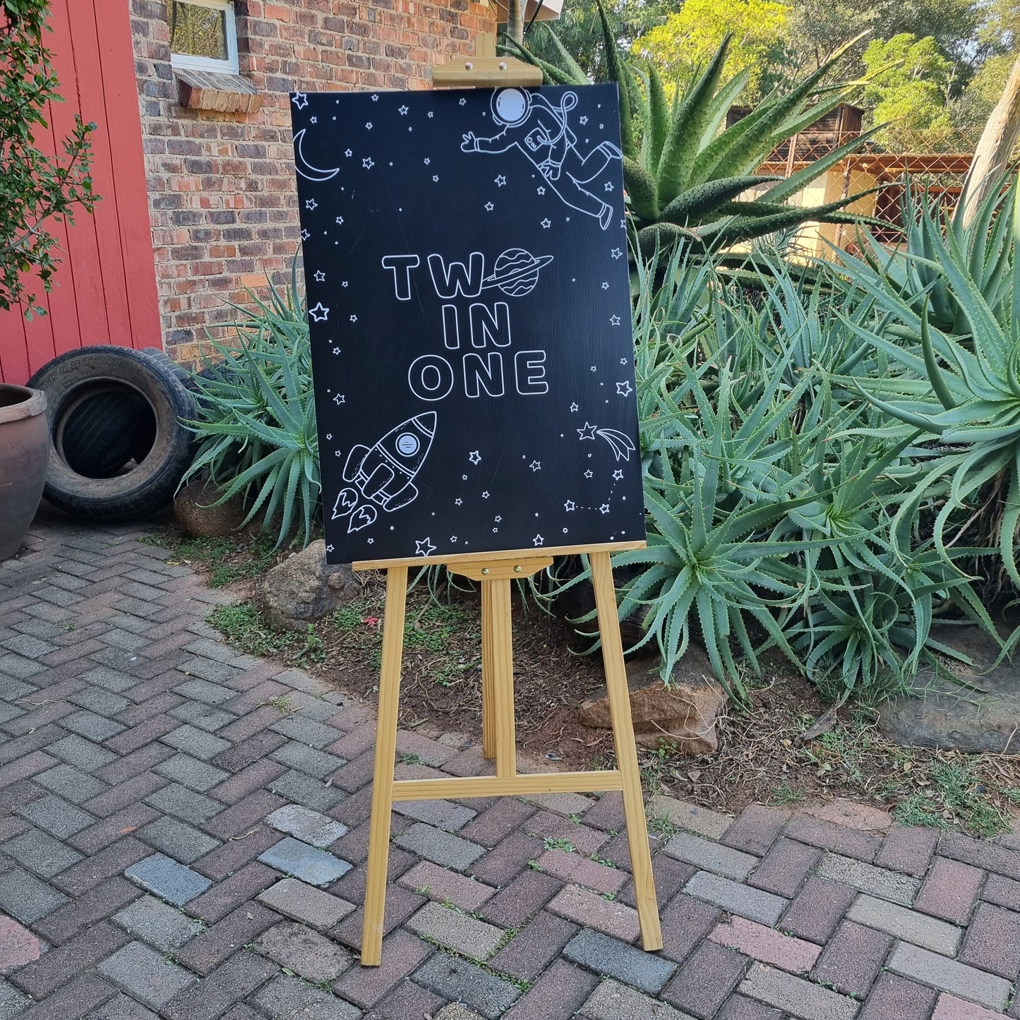 Black sign with 'Two in One' text on a stand outdoors next to plants