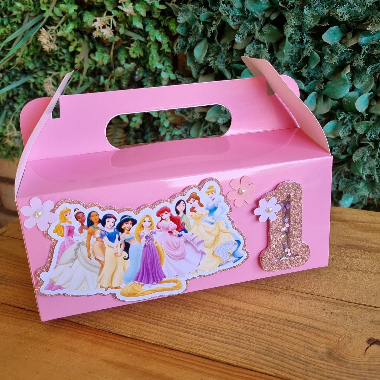 Pink children's lunch box with Disney Princess design on a wooden surface with greenery in the background