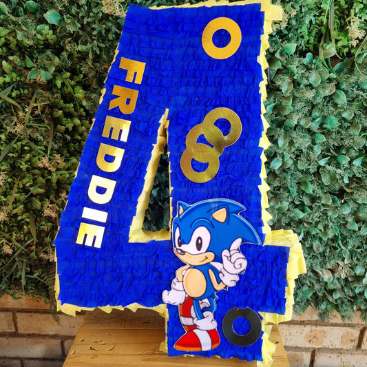 A large blue and yellow piñata shaped like the number 4, featuring Sonic the Hedgehog graphics and the name 'FREDDIE' in gold letters, displayed on a wooden table in front of a green hedge background.