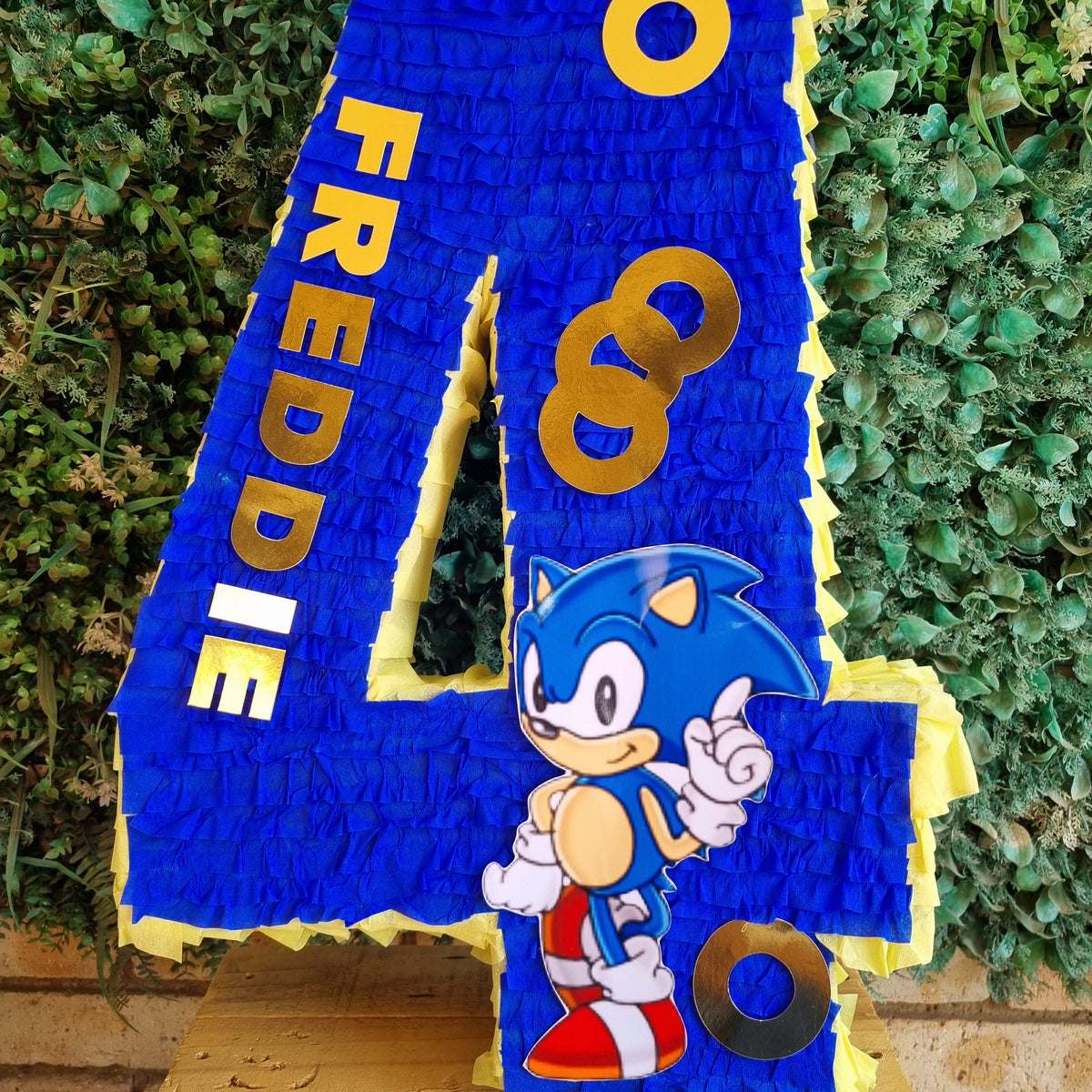 A large blue piñata shaped like the number 4, decorated with Sonic the Hedgehog artwork and the name 'FREDDIE' in gold letters, displayed on a wooden surface in front of a green hedge background.