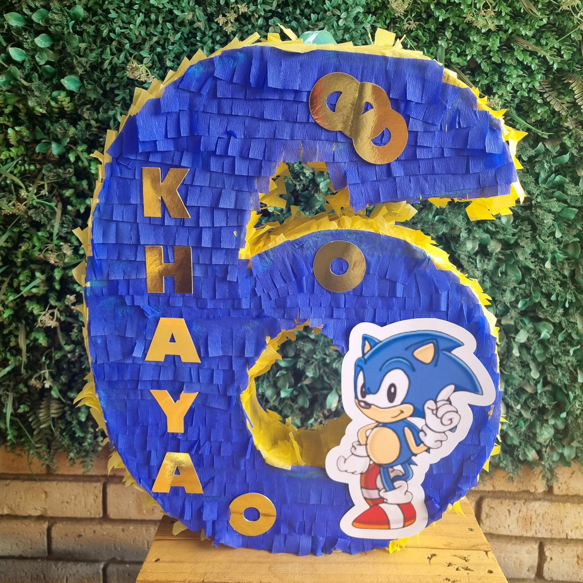 A large blue and yellow shredded paper piñata shaped like the number 6, with the name 'KHAYO' in gold letters and a Sonic the Hedgehog character cutout, displayed on a wooden table in front of a green hedge wall.