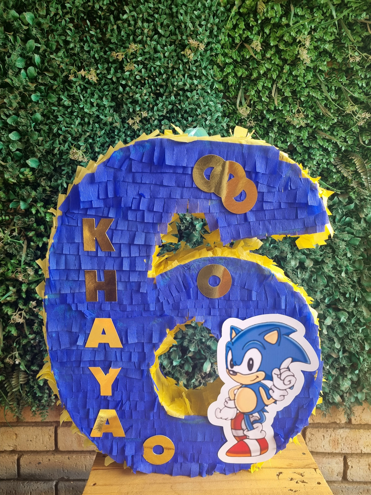 A custom blue and yellow 6th birthday piñata shaped like the number 6, featuring the name 'KHAYAO' in gold, gold '60' balloons, a Sonic the Hedgehog character sticker, and green leafy accents, displayed against a green hedge background.