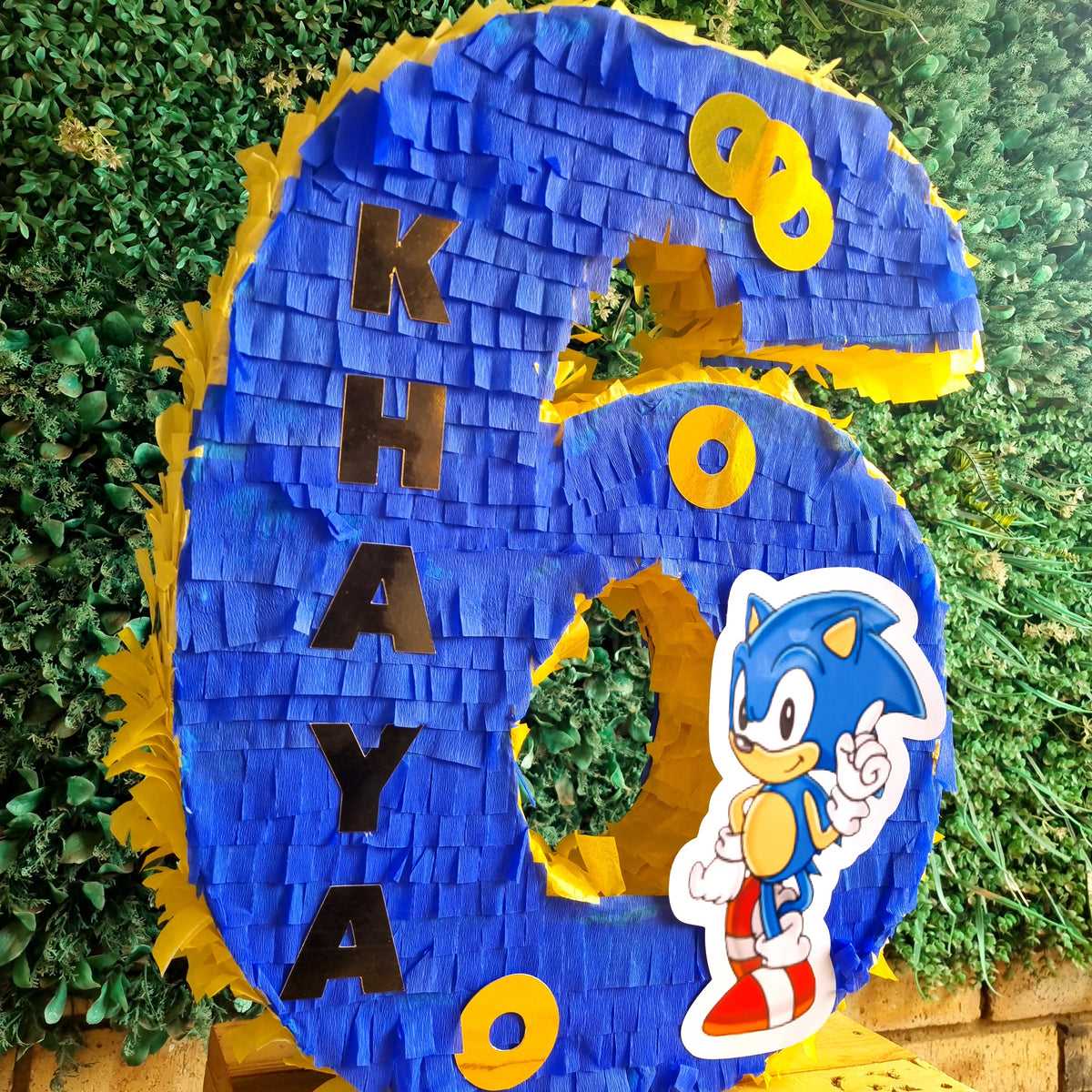 A large blue and yellow piñata shaped like the number 6, personalized with the name 'KHAYA' in black letters and featuring a Sonic the Hedgehog character cutout. The piñata is displayed on a wooden surface in front of a green hedge background.