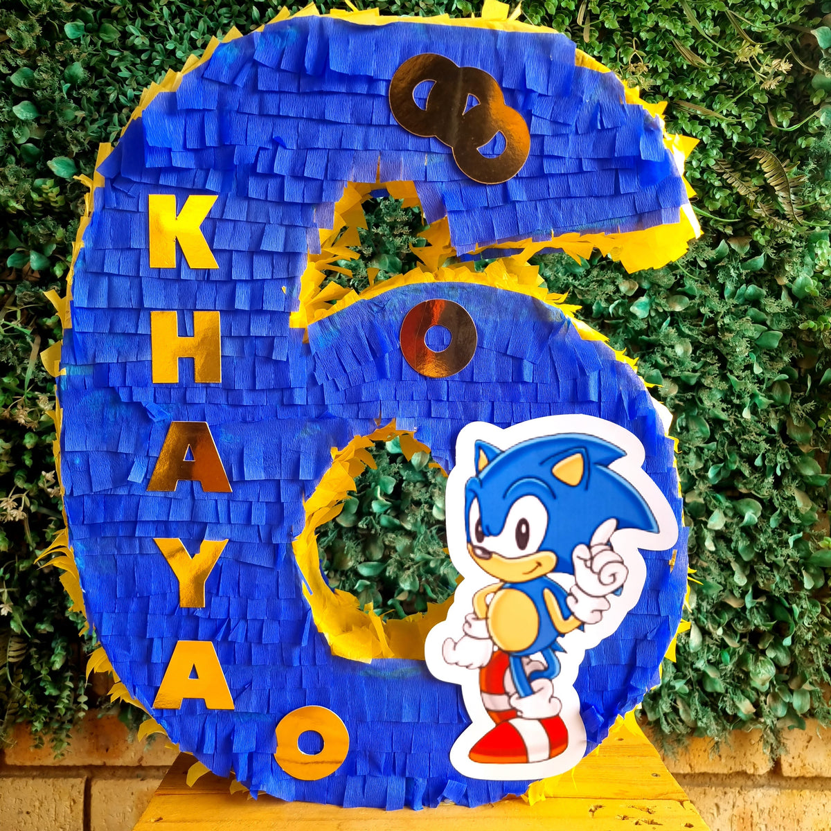 A custom blue and yellow 6th birthday piñata shaped like the number 6, featuring the name 'KHAYA' in yellow letters and a Sonic the Hedgehog character graphic. The piñata is displayed on a wooden table in front of a green hedge background.