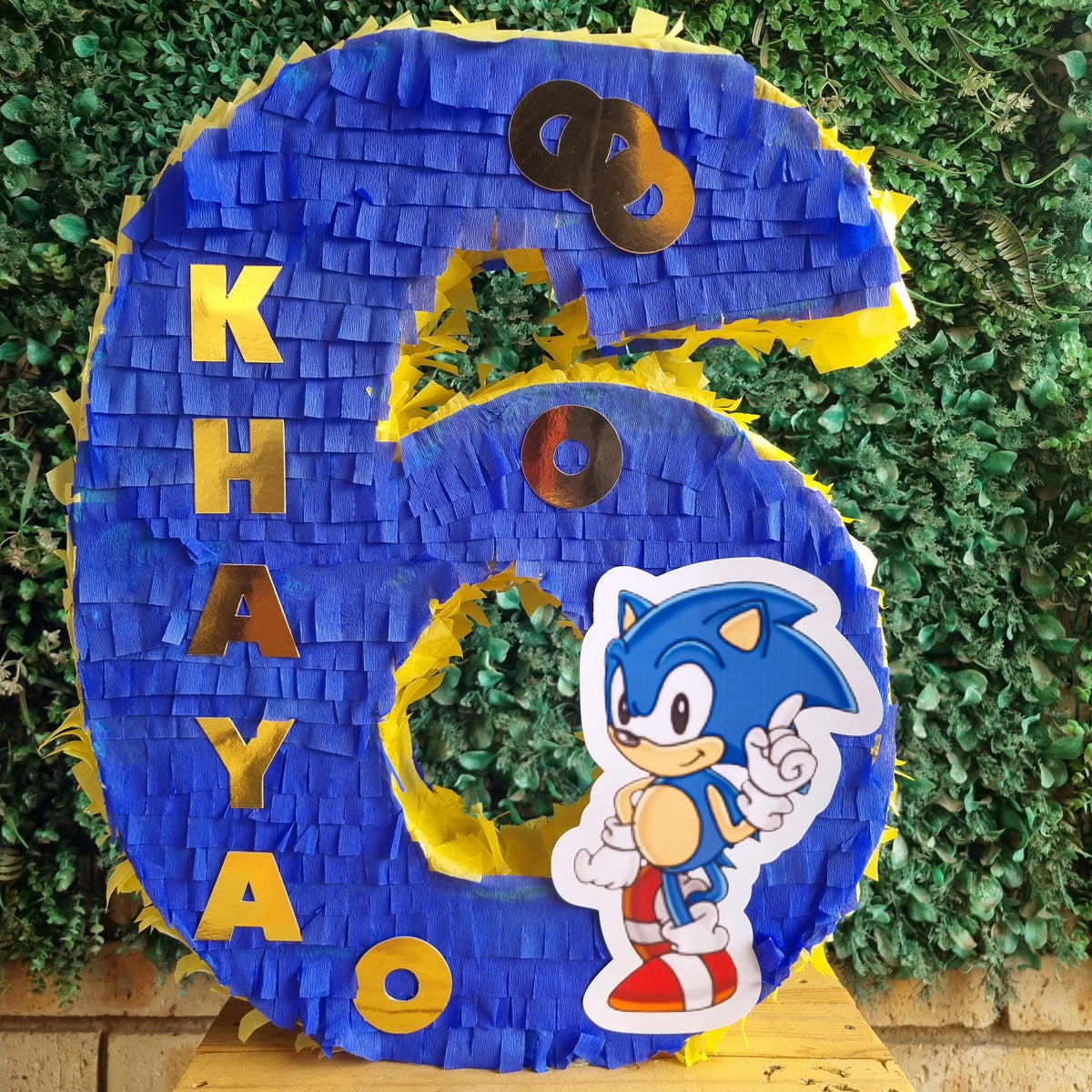 A large blue and yellow piñata shaped like the number 6, decorated with the name 'KHAYA' in gold letters, a Sonic the Hedgehog character sticker, and metallic gold number 8 accents, displayed on a wooden table against a green hedge background.
