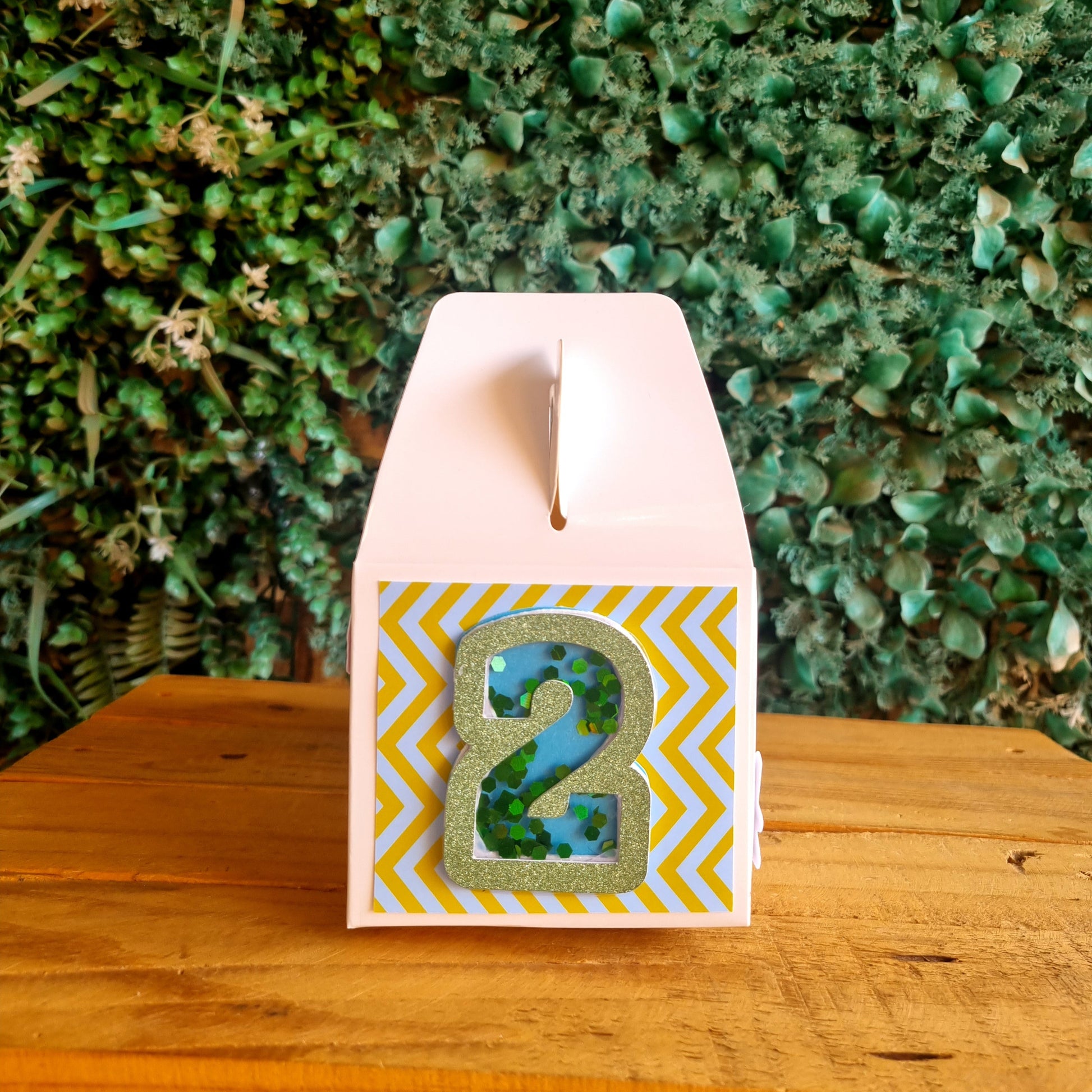 Decorative box with a green design on a yellow and white chevron pattern, attached to a wooden block against a green leafy background.