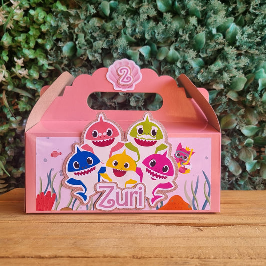 Pink gift box with cartoon fish design on a wooden surface and green plants in the background