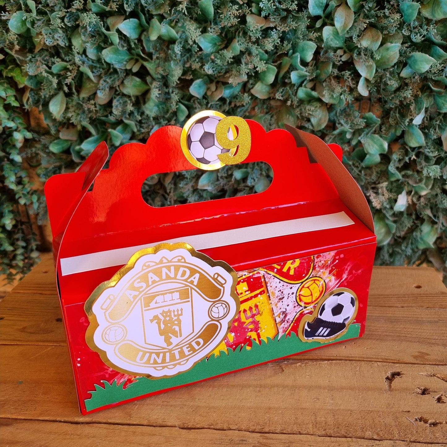 Red decorative item with gold and white design on a wooden surface and green foliage background