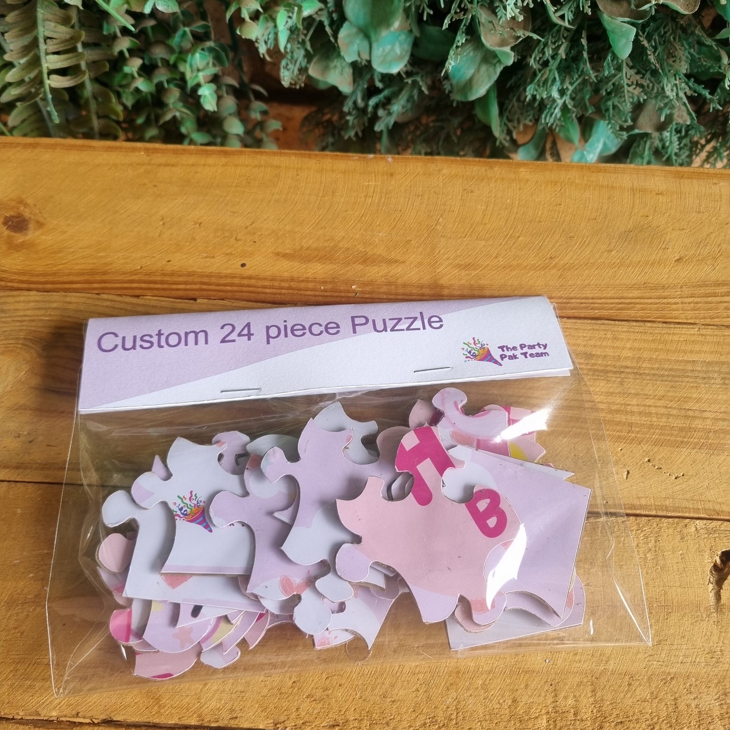 Pink and white puzzle pieces in clear packaging on a wooden surface with greenery.