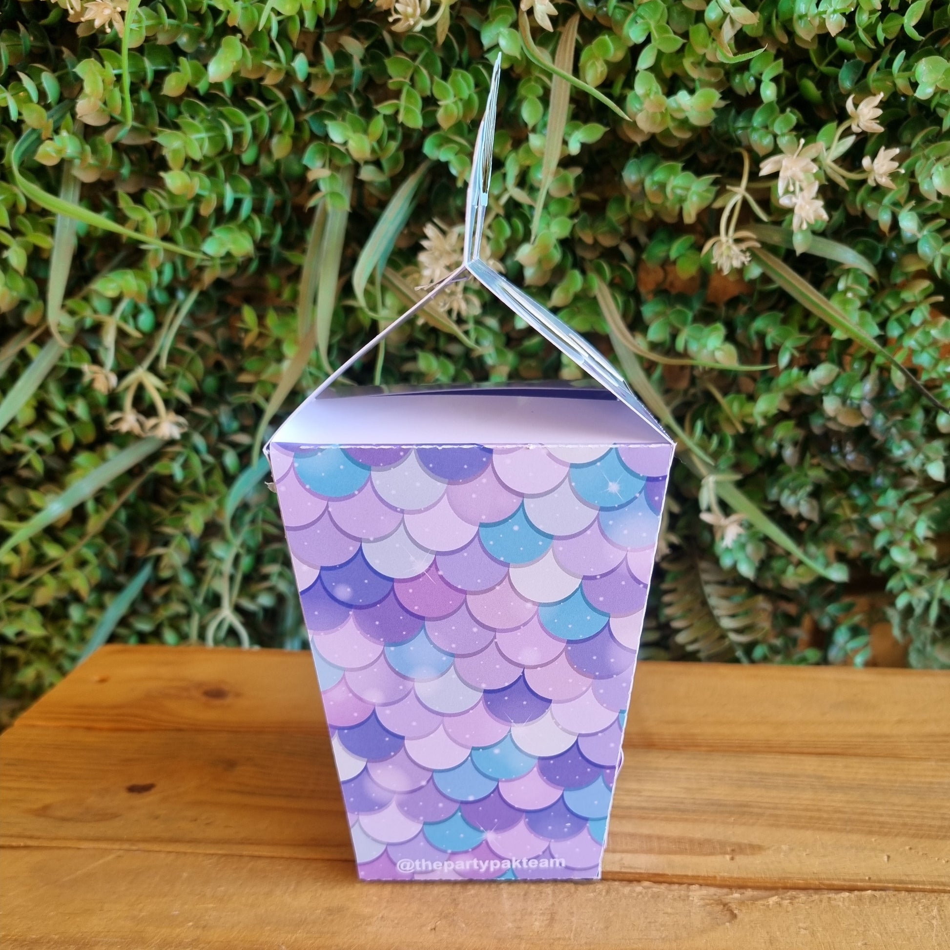 Small purple box with a fish scale pattern on a wooden surface with greenery in the background