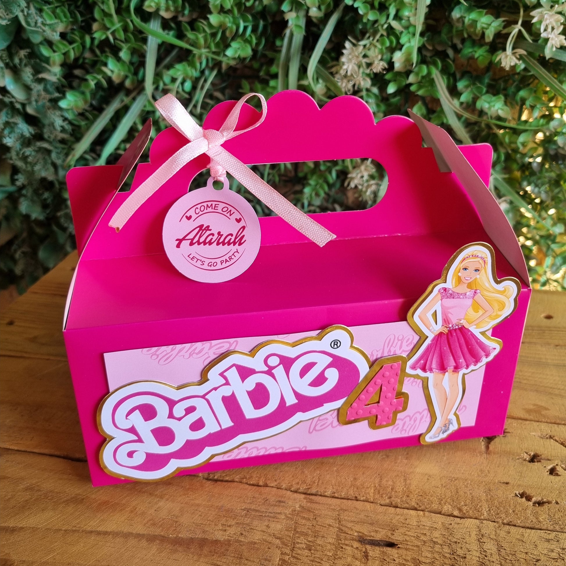 Pink Barbie toy box with a bow on a wooden surface and greenery background