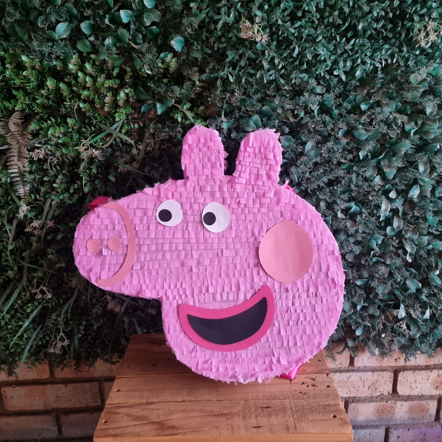 Pink pig-shaped pinata against a green bush and wooden crate background