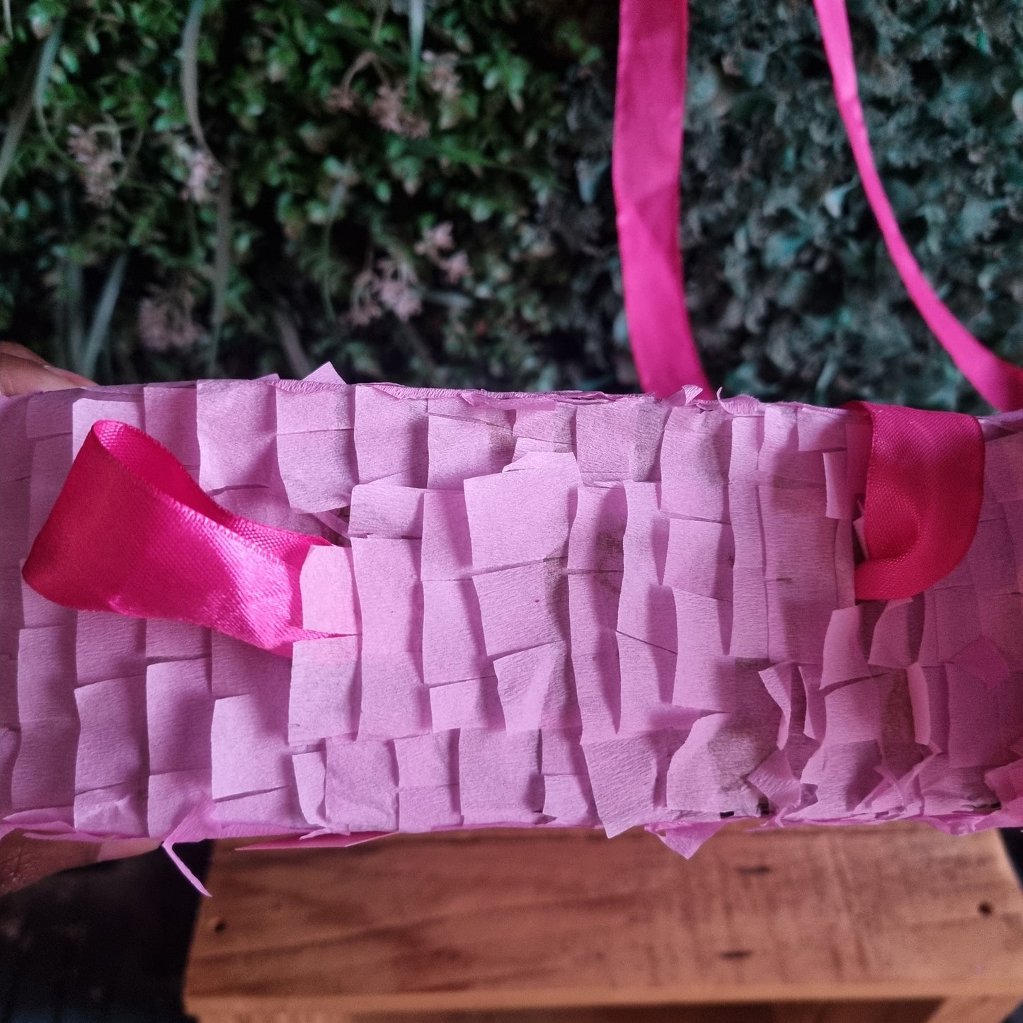 Pink piñata with ribbons against a green bush and wooden crate background