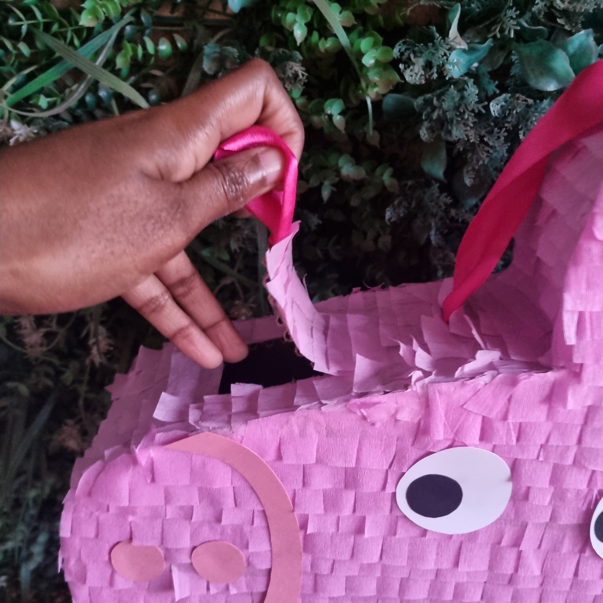 Pink piñata shaped like a horse with a hand holding a pink ribbon against a green leafy background