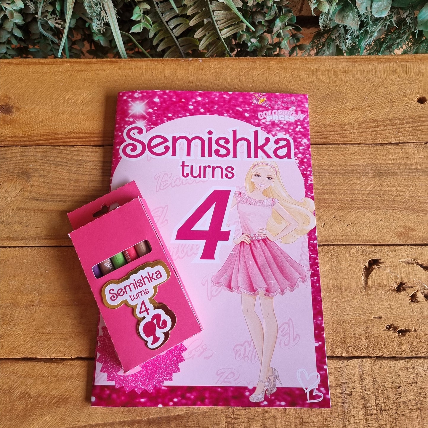 Pink birthday card with 'Semishka turns 4' on a wooden surface with greenery.