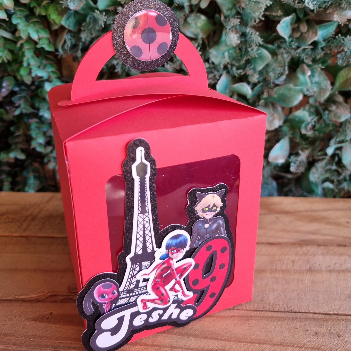 Red gift box with decorative handle featuring a design on a wooden surface with greenery.