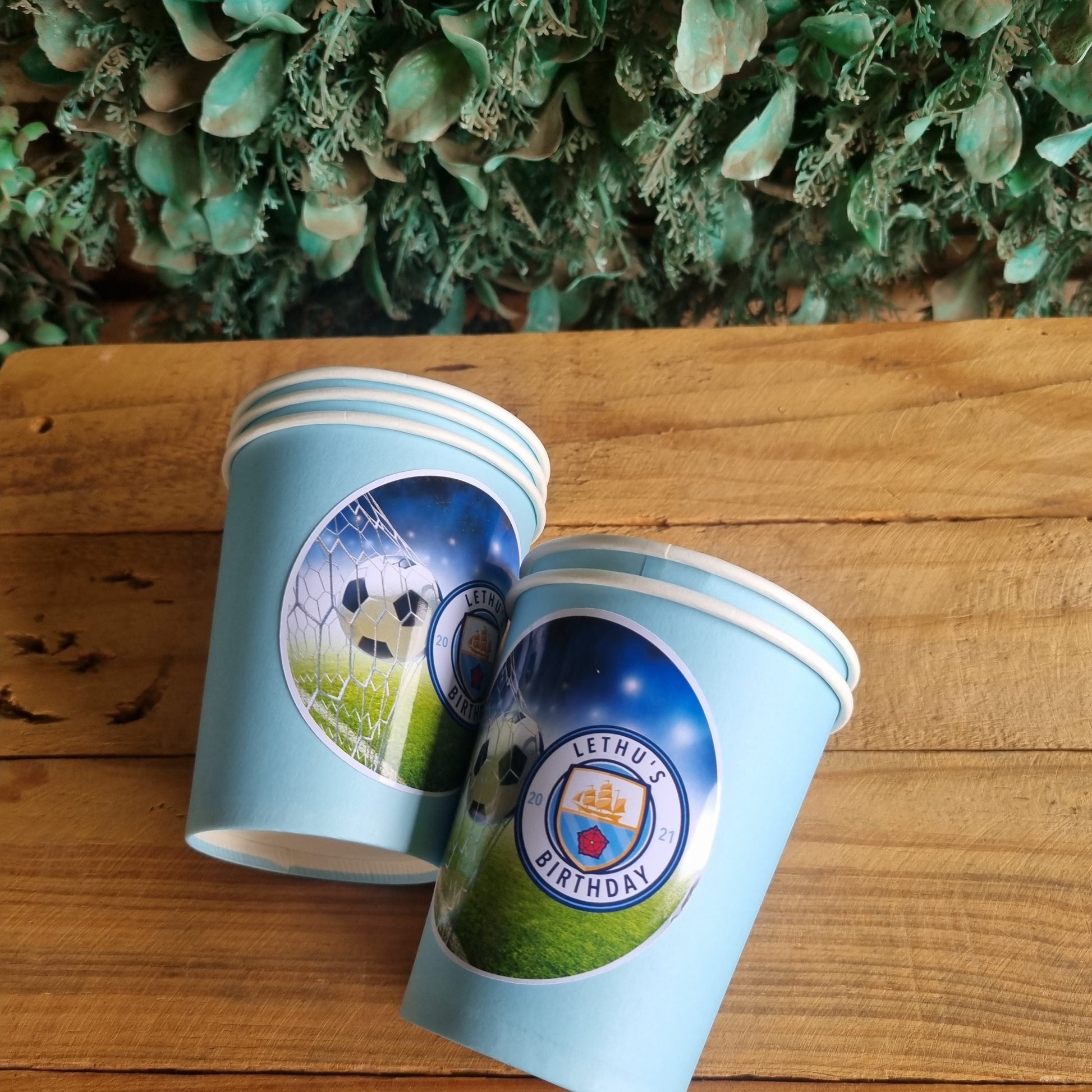 Two paper cups with festive designs on a wooden surface with a green leafy background