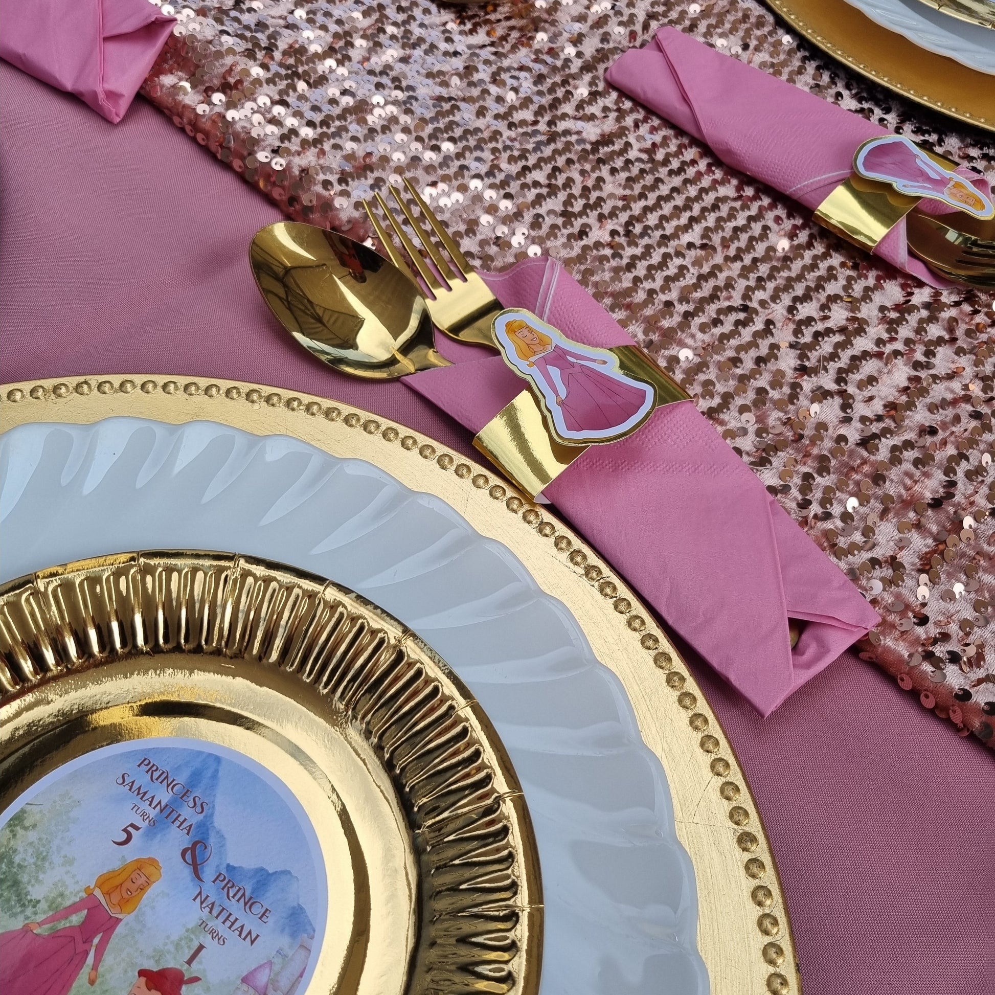 Decorative table setting with gold-rimmed plates, pink napkins, and fairy tale-themed cutlery on a pink sequined tablecloth.