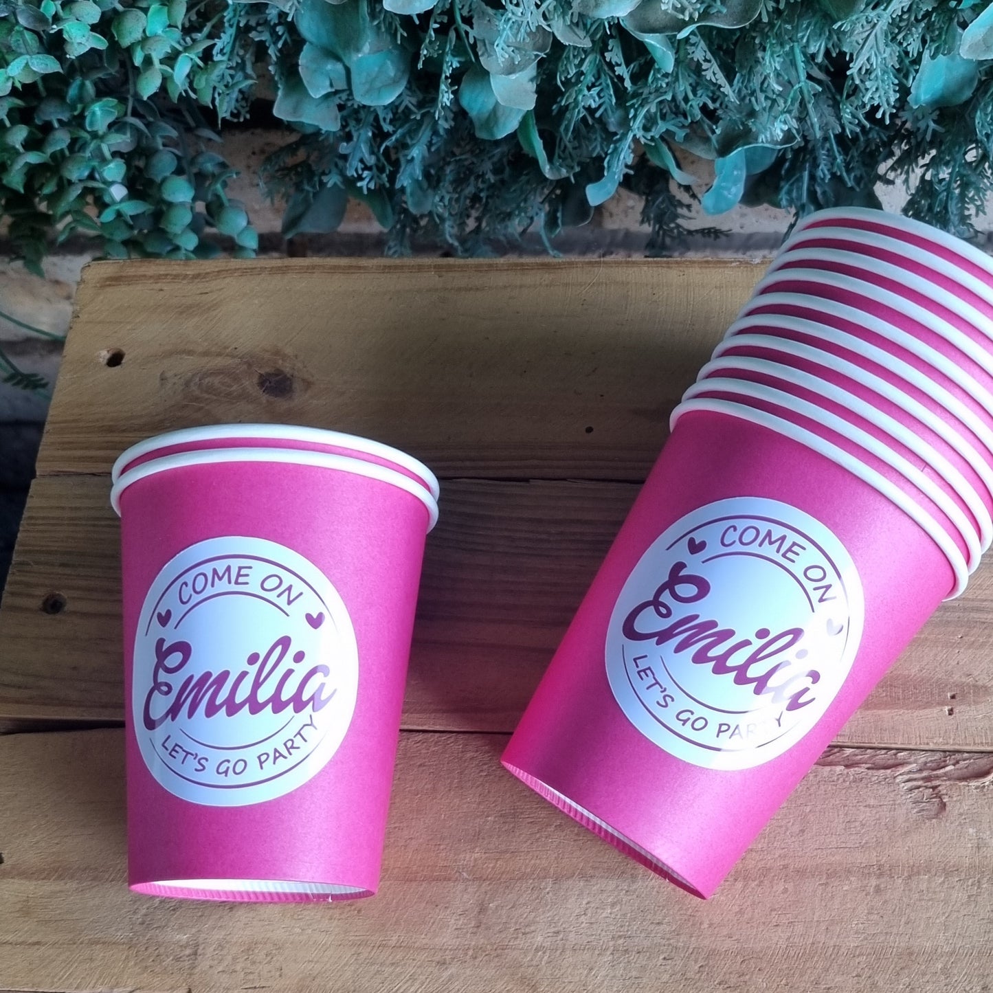 Two pink cups with a logo on a wooden surface next to green foliage.