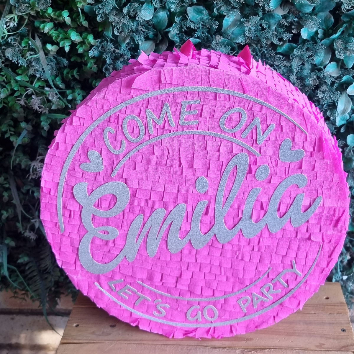 A round pink woven party  Barbie piñata with silver glitter text reading 'COME ON Emilia LET'S GO PARTY', displayed on a wooden surface against a green leafy background.