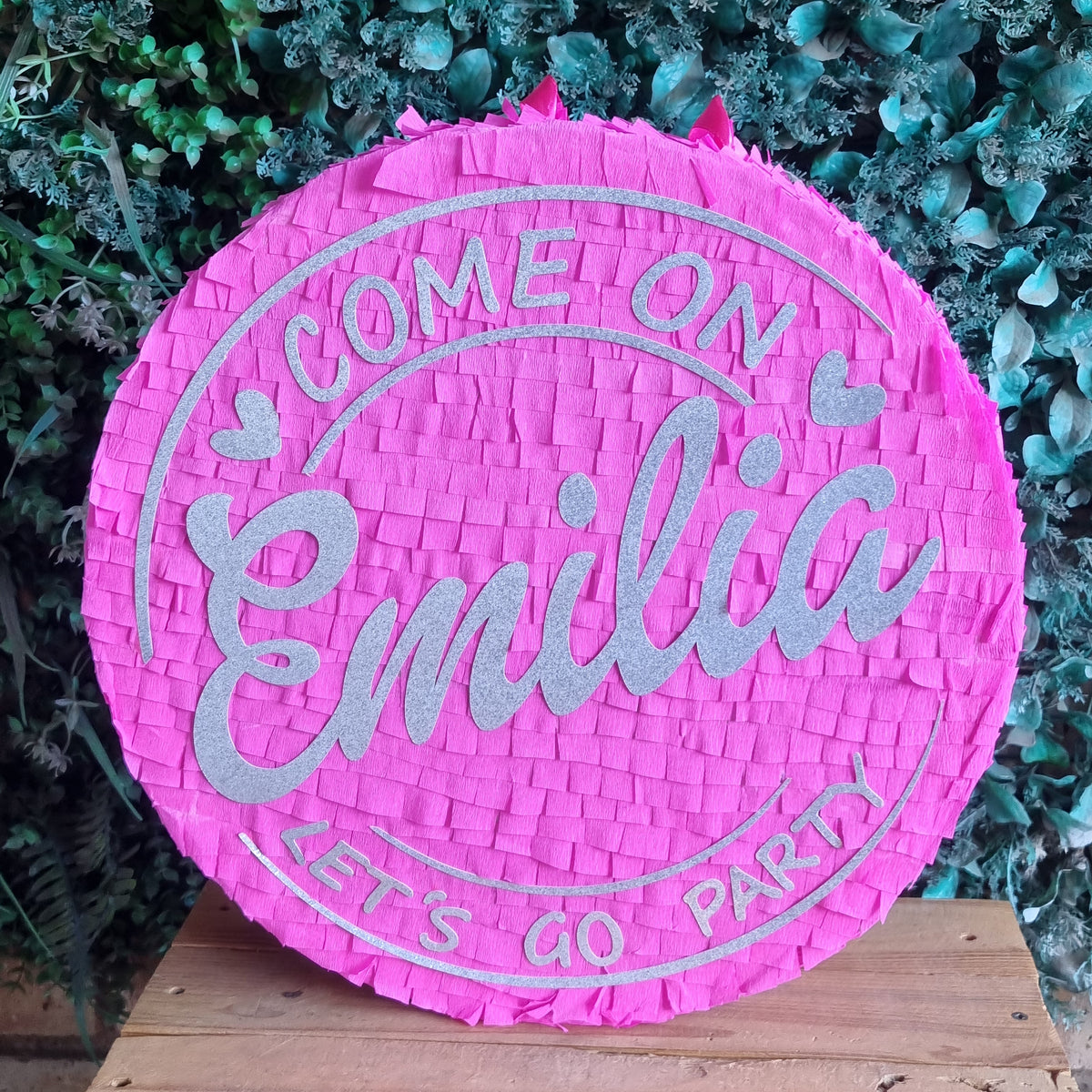 A round pink woven paper Barbie piñata with silver glitter text reading 'COME ON Emilia LETS GO PARTY', displayed on a wooden surface against a green leafy background