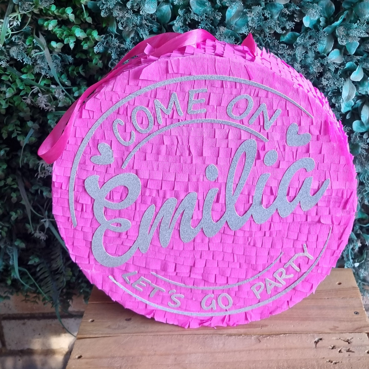 A round pink woven  Barbie pinata with silver glitter text reading 'COME ON Emilia LET'S GO PARTY', displayed on a wooden surface against a green leafy background.