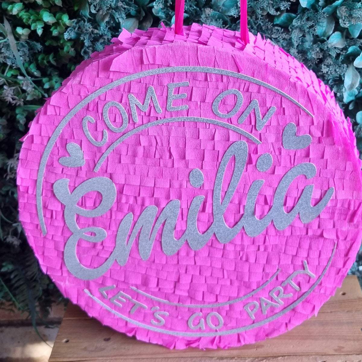 A round pink woven party  Barbie piñata with silver glitter text reading 'COME ON Emilia LET'S GO PARTY', hanging against a green leafy backdrop.