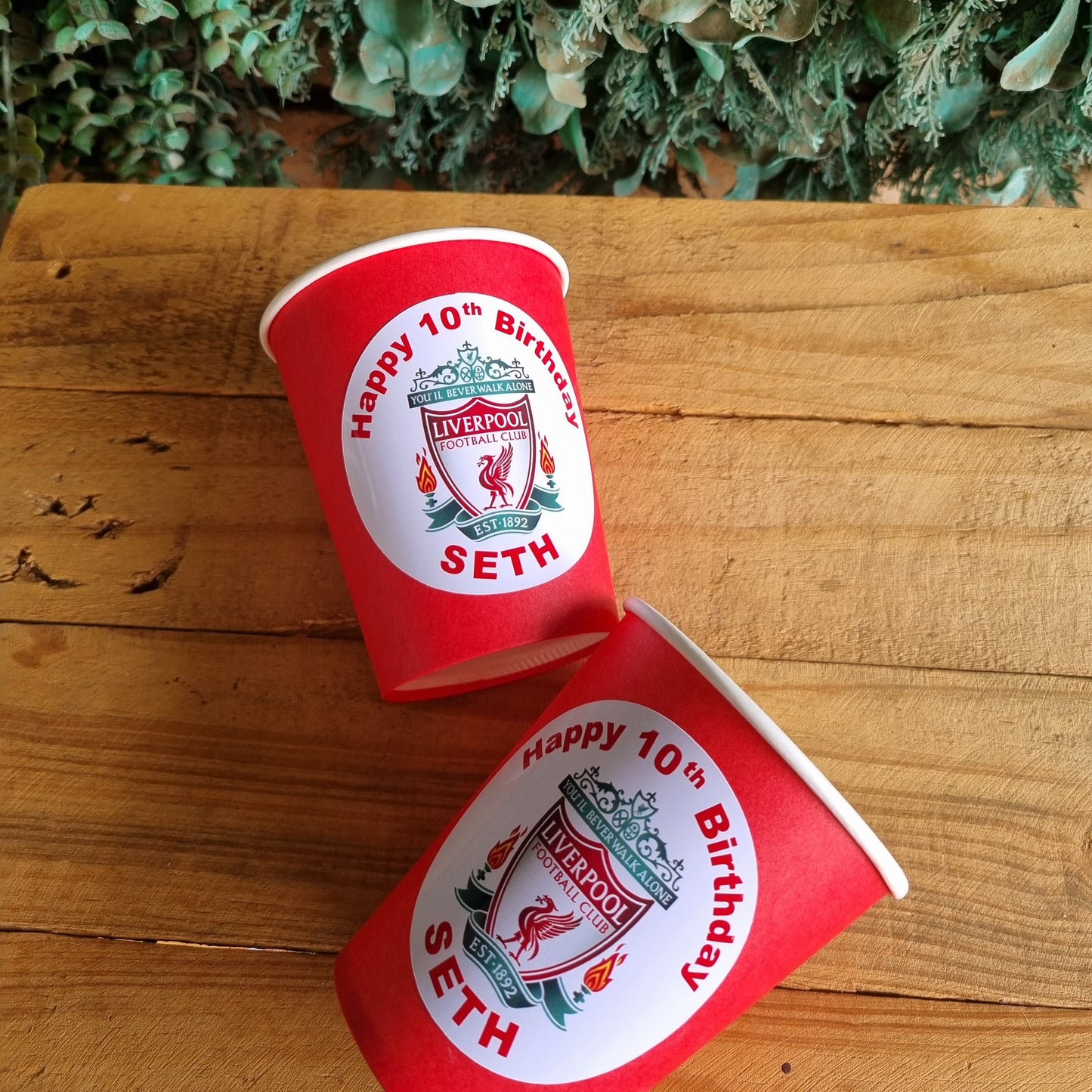 Two red paper cups with white labels on a wooden surface next to green foliage.