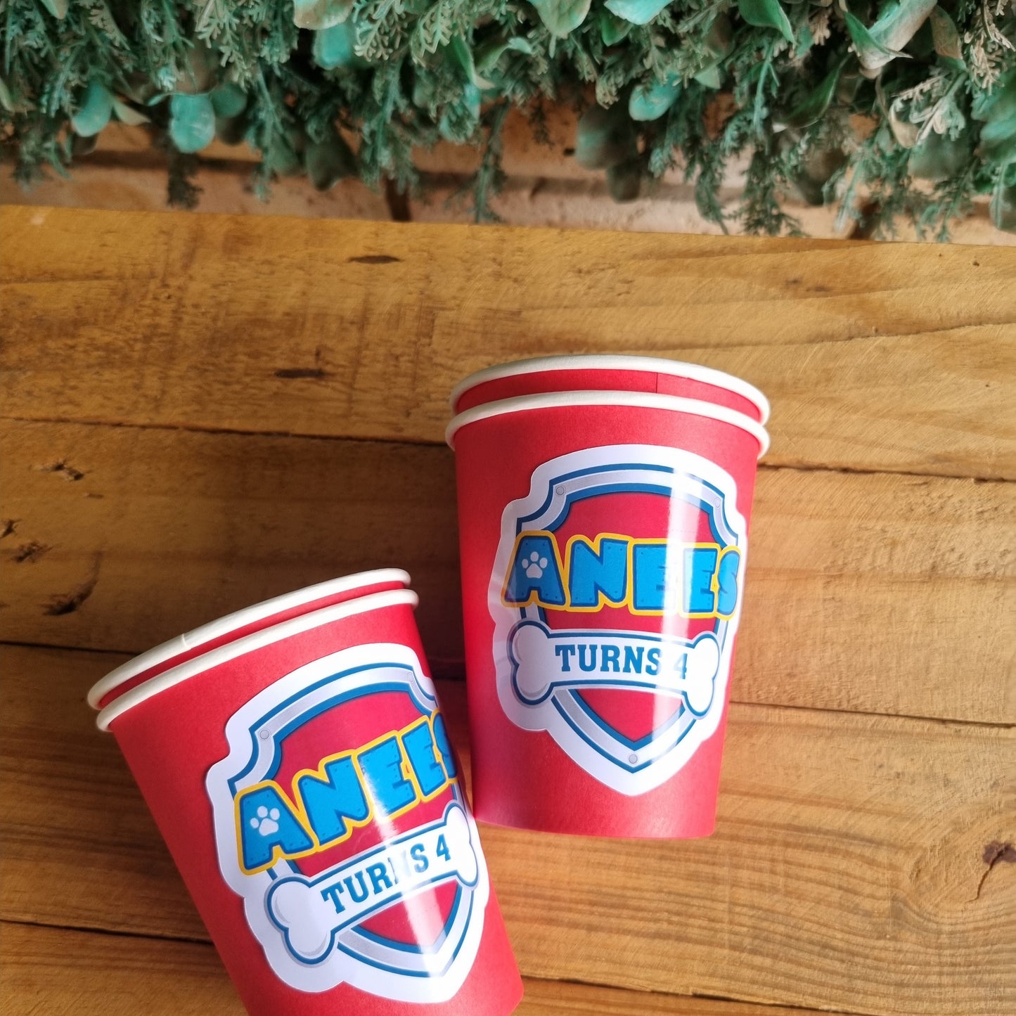 Two red paper cups with a logo on a wooden surface next to greenery.