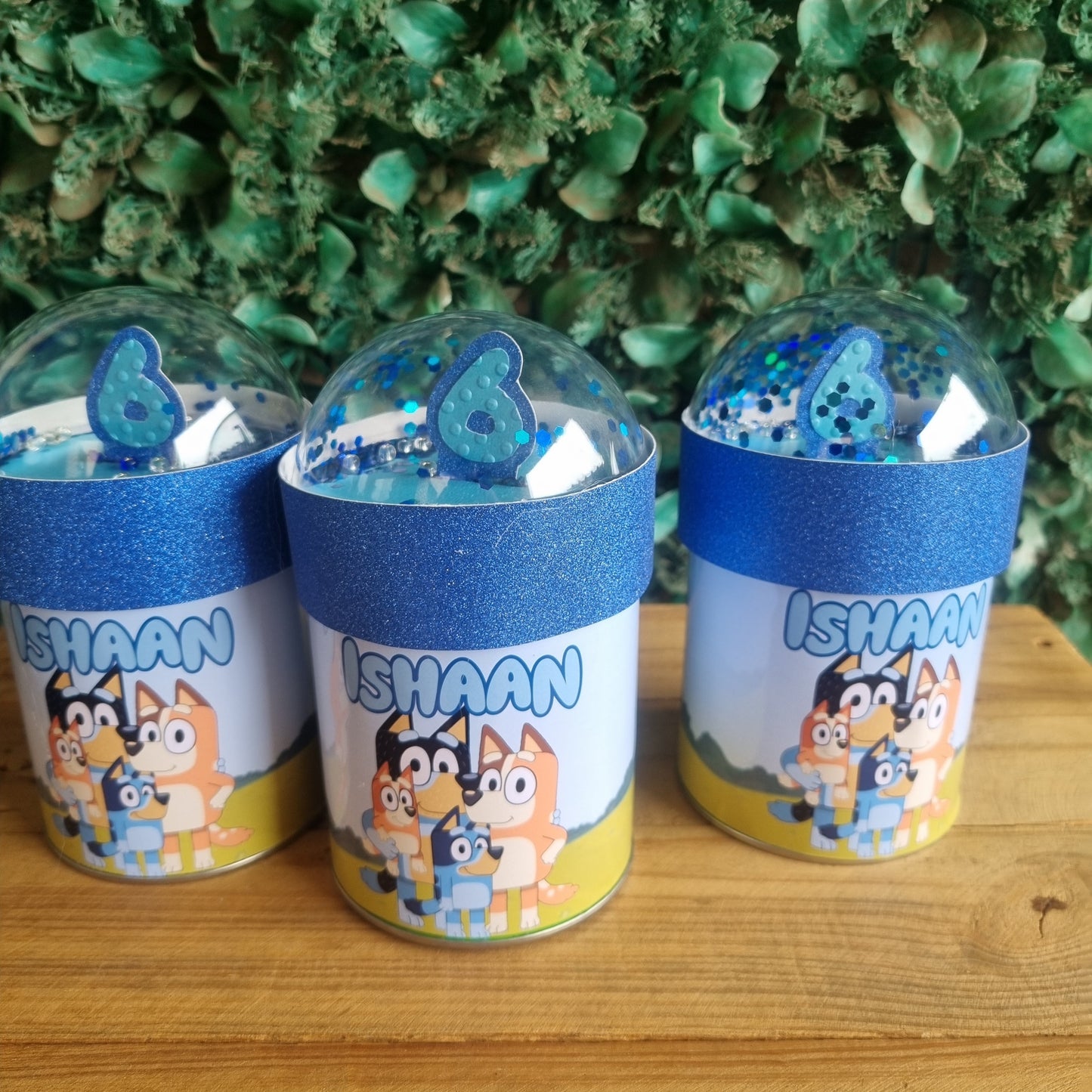 Three cylindrical containers with cartoon character design on a wooden surface and green foliage background