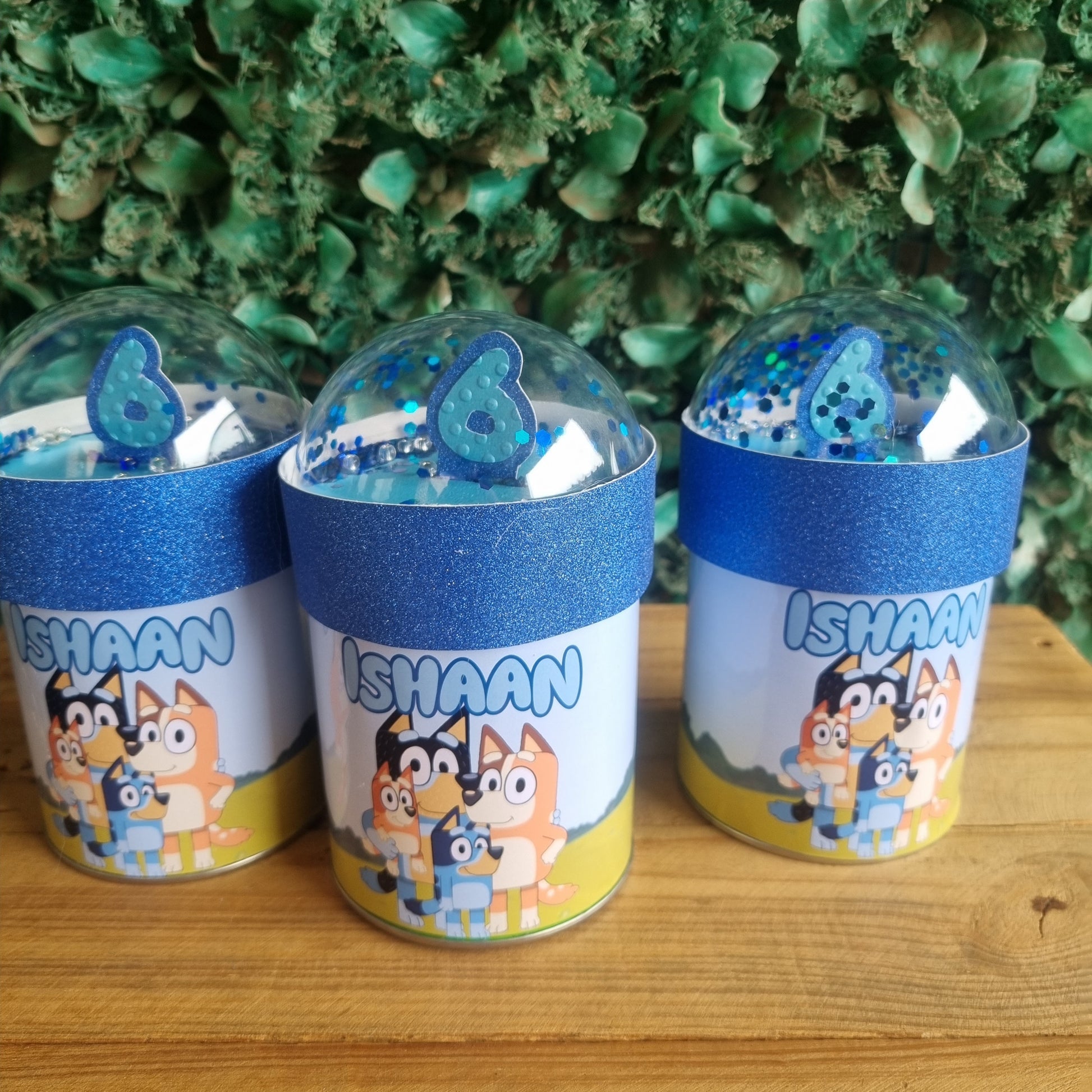 Three cylindrical containers with cartoon character design on a wooden surface and green foliage background