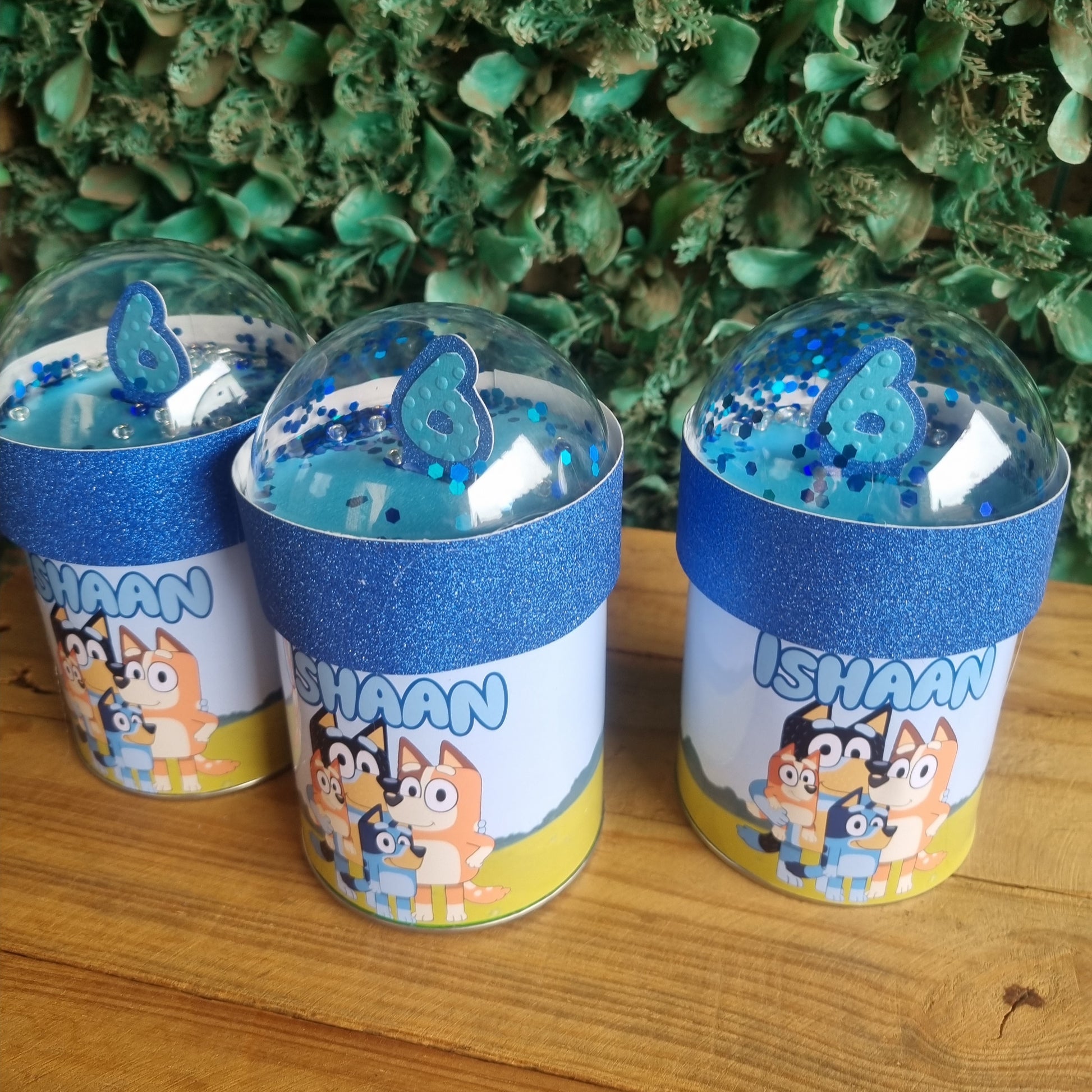 Three cylindrical containers with blue lids on a wooden surface next to green foliage.