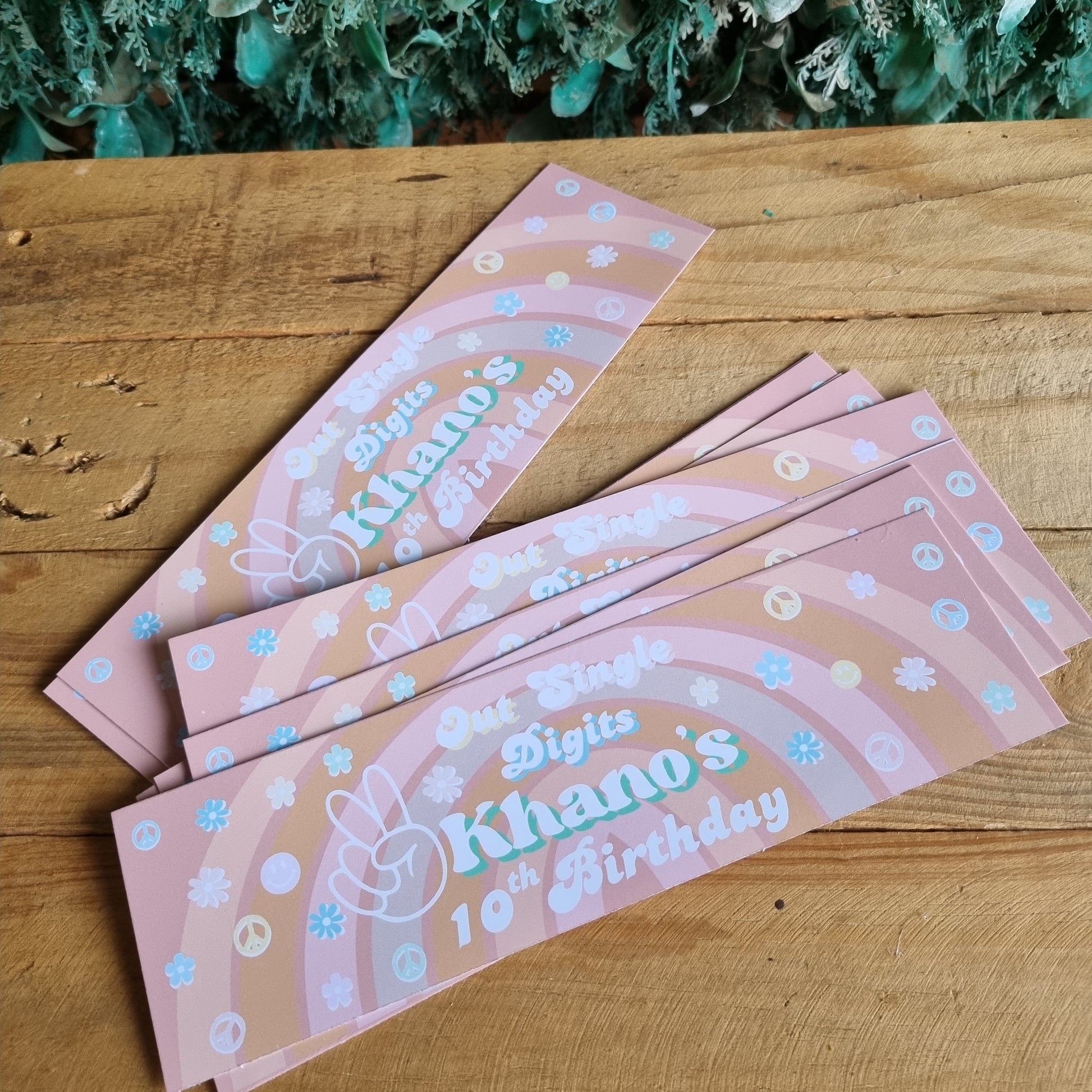 Pink bookmarks with decorative text and designs on a wooden surface next to green foliage.
