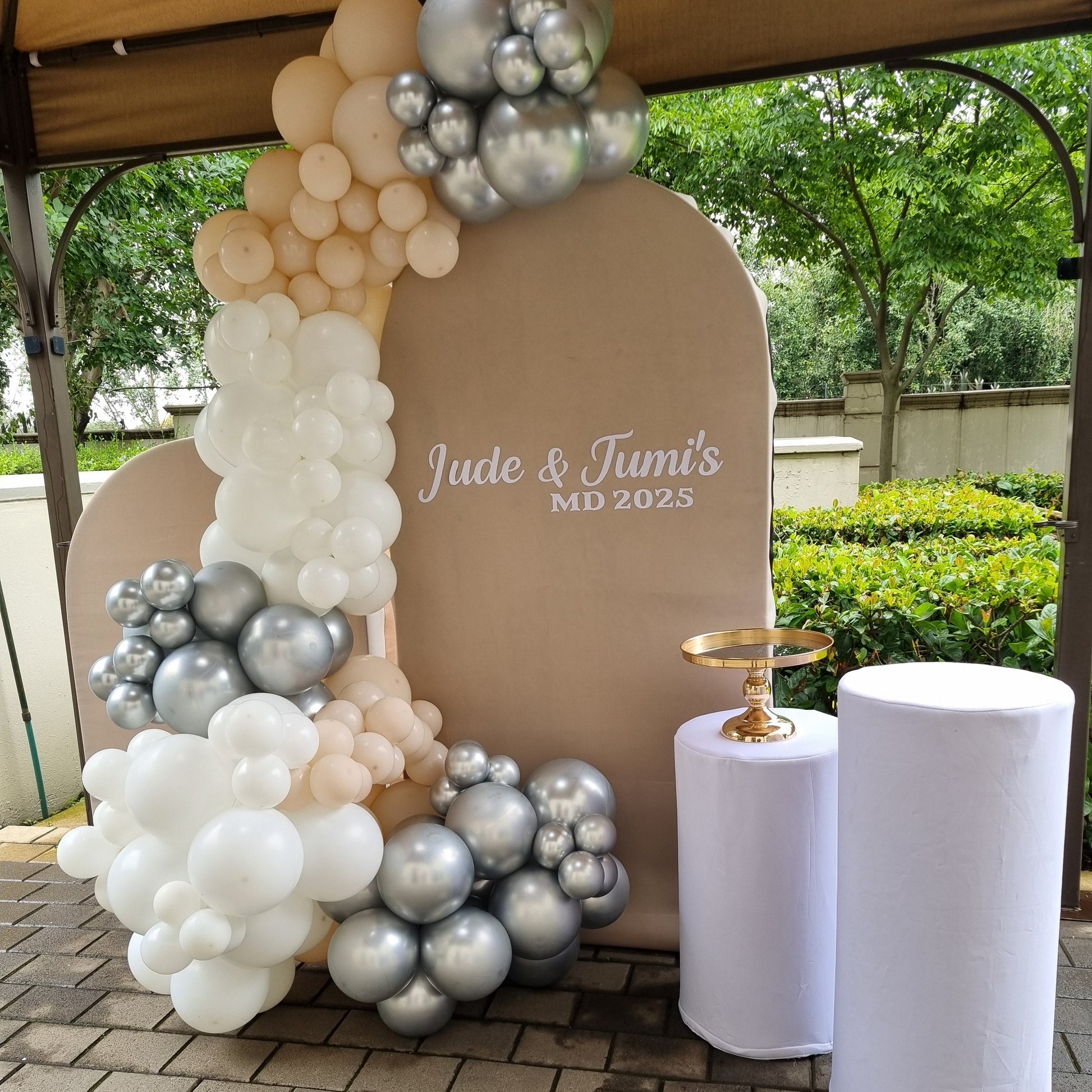 Decorative arch with balloons and a sign reading 'Jude & Tamis MD 2025' on a tiled floor.